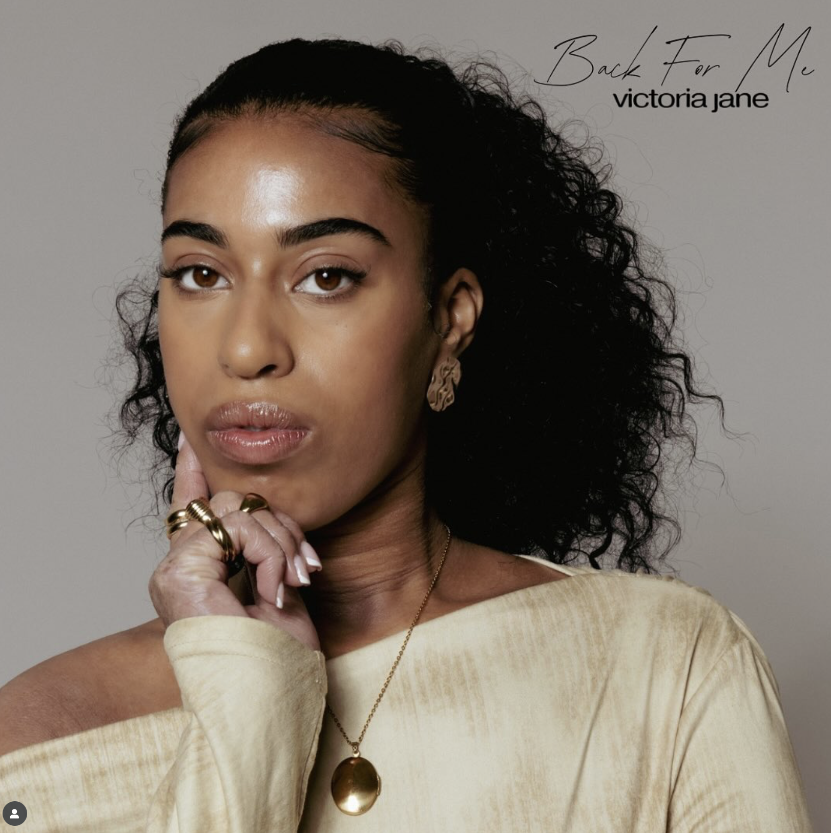 A woman with dark curly hair, wearing gold jewelry and a beige top, posing with her hand resting on her chin. Professional beauty shoot inside Manchester photography studio – The Space MCR.