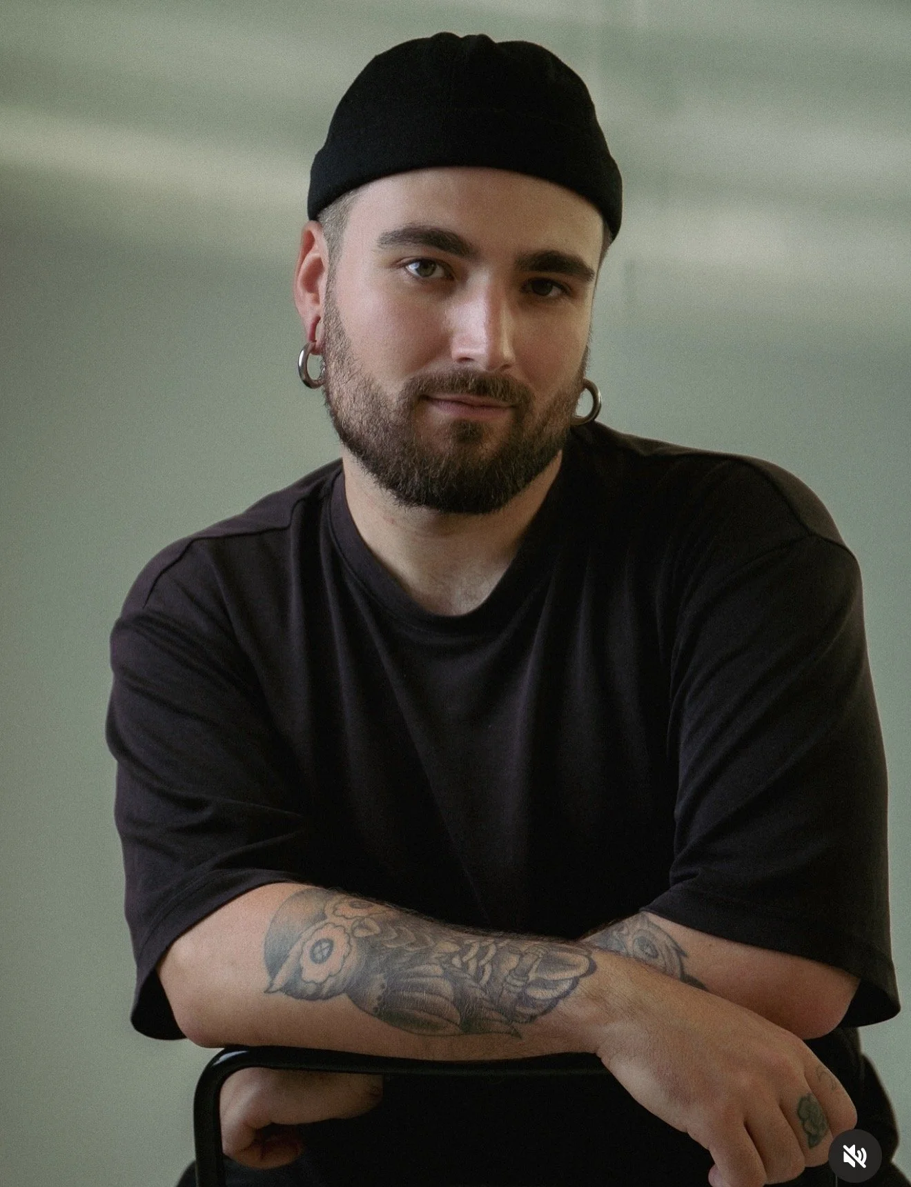 Young man with beard, earrings, and tattoos on his arm, wearing a black beanie and black shirt, sitting with arms crossed, looking at the camera.
