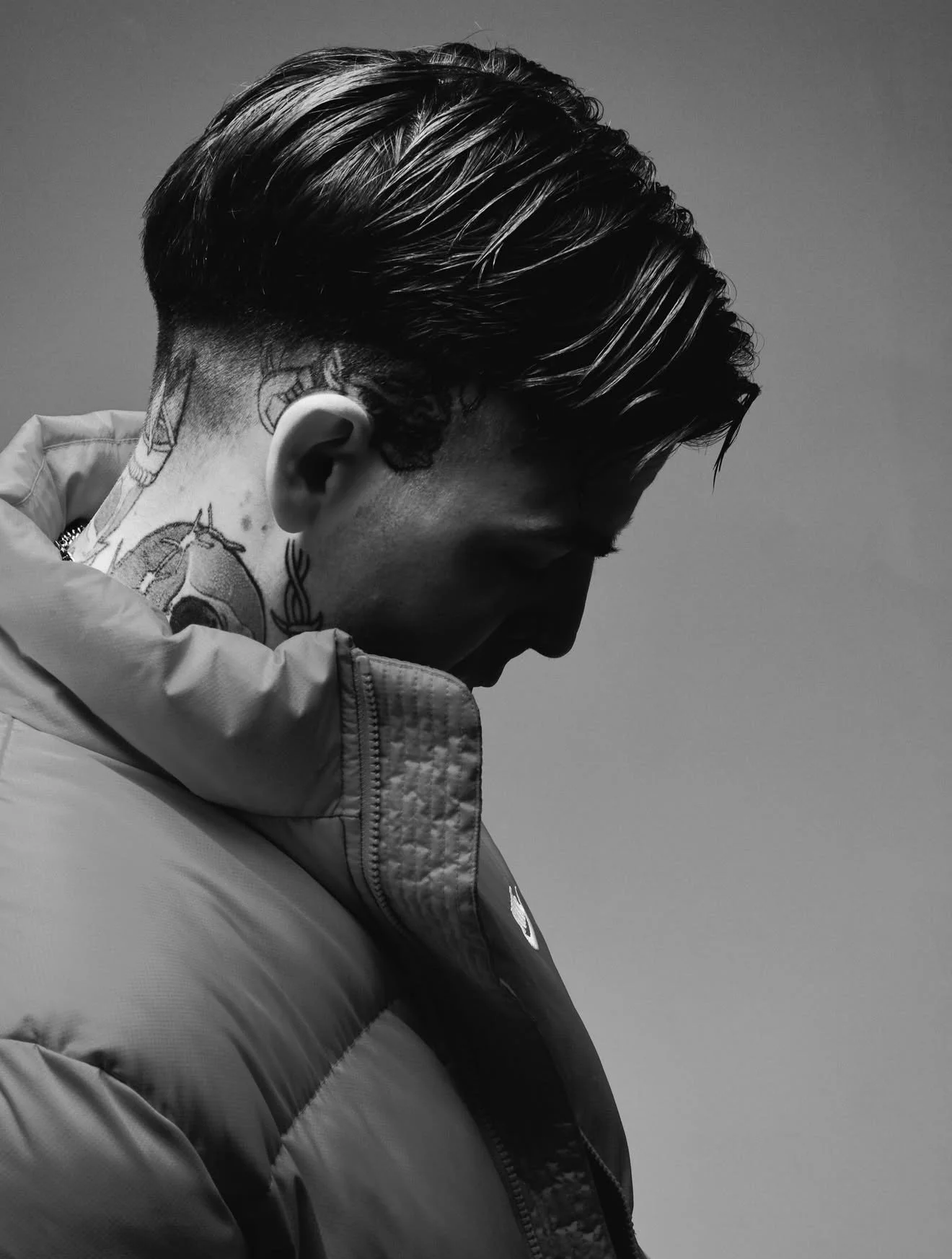 Black and white photo of a man with styled hair, tattoos on his neck, and wearing a puffy jacket, looking down.