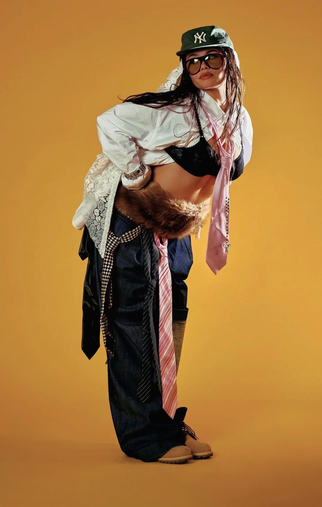 Woman dressed in a mix of casual and eclectic fashion, wearing a green baseball cap with a New York Yankees logo, large glasses, a white jacket, a black lace bralette, a pink tie, baggy dark jeans with added accessories, and hiking boots, with a yell