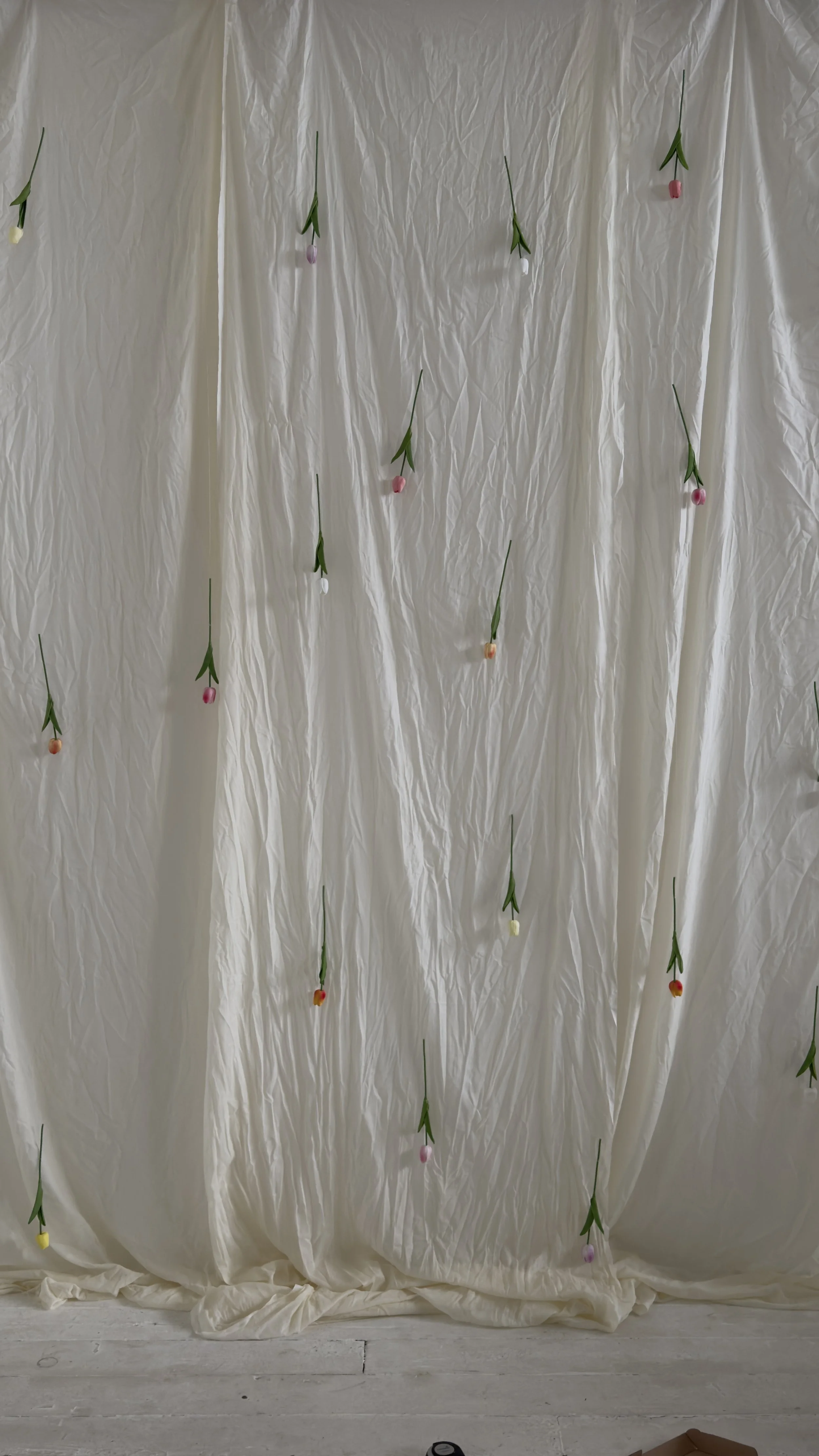 A white fabric backdrop with small, colorful, artificial flowers attached, creating a simple decorative display.
