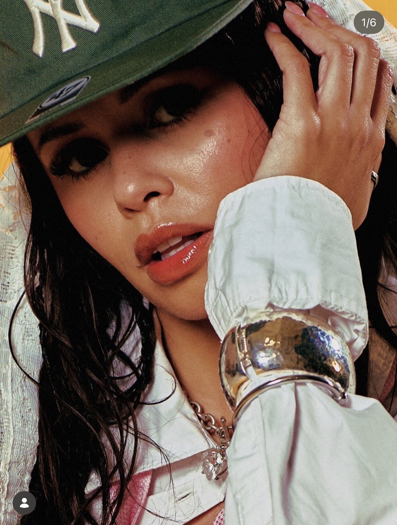 Close-up of a woman wearing a green New York Yankees cap, white jacket, and jewelry, resting her head on her hand with wet, wavy black hair, and glossy lips.