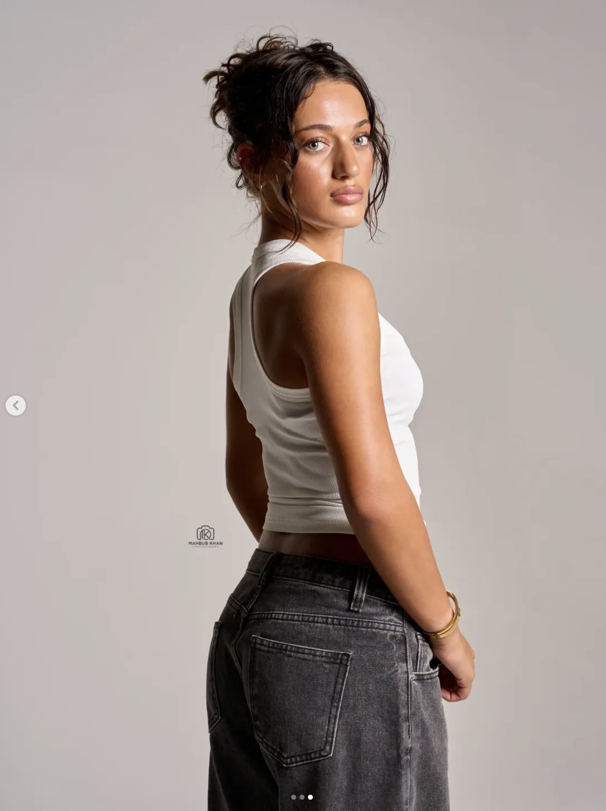A young woman with wavy brown hair, wearing a white sleeveless top and grey jeans, looking over her shoulder against a plain light gray background. Lifestyle photoshoot setup at The Space MCR photography studio in Manchester.