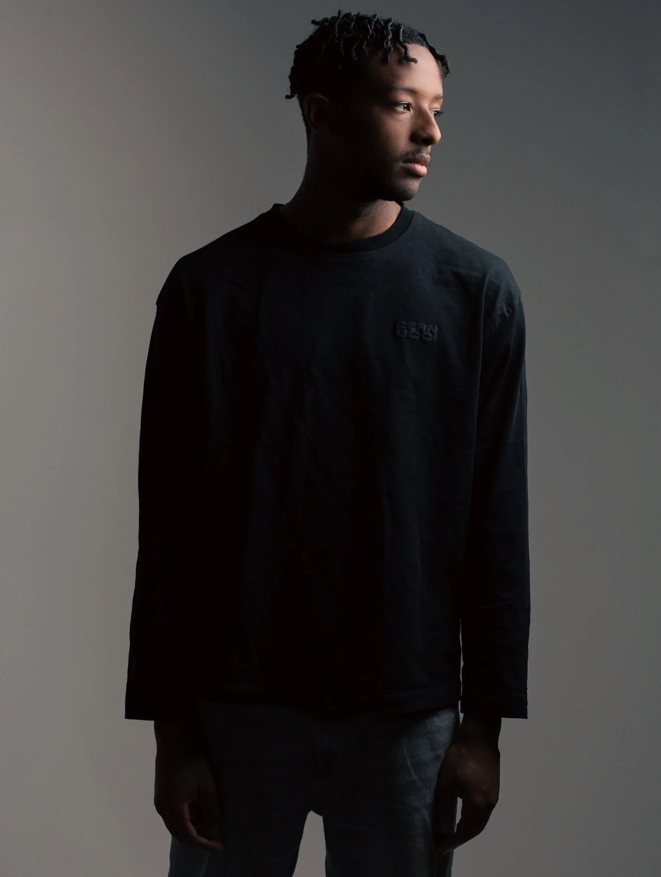 A young man with dark skin and short dreadlocks standing against a neutral background, looking to his right. He is wearing a long-sleeved dark shirt and light-colored pants.