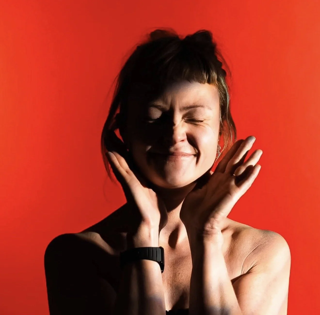 A woman with short brown hair and a black watch on her left wrist, smiling with her eyes closed and hands gently touching her face, against a vibrant red background.