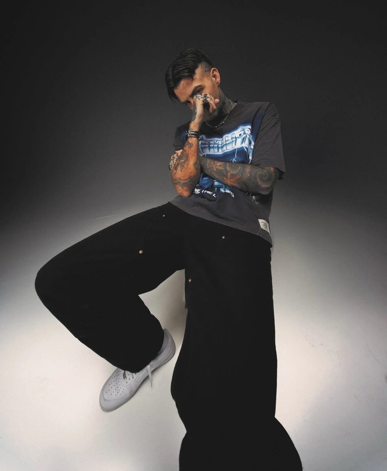 A young man with tattoos, dark hair, and wearing a black graphic t-shirt, black pants, and white sneakers, poses against a dark gray background with one leg raised and hand covering part of his face.