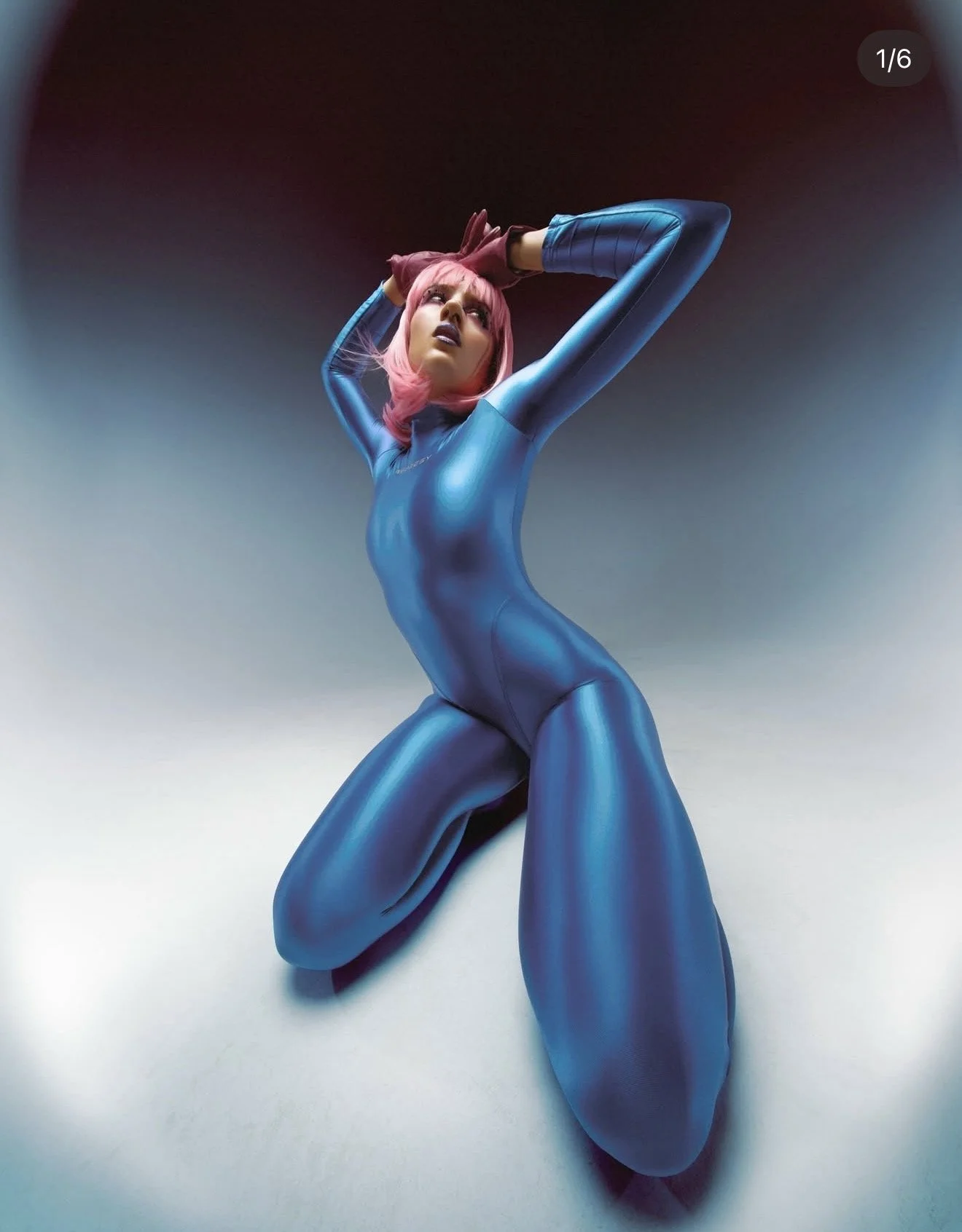 Person dressed in a shiny blue bodysuit with pink hair, posing with arms raised above head against a gradient background.