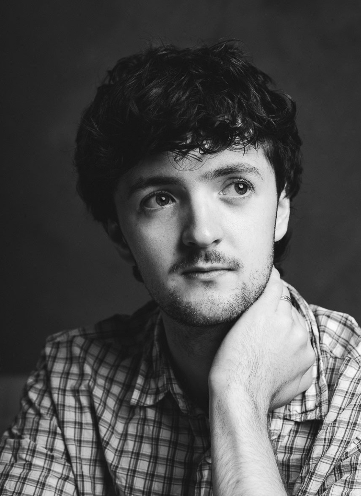 Young man wearing a checkered shirt, looking slightly upward to the right, with his right hand resting on his neck, in black and white portrait photography. Creative corner and textured photo backdrop inside Manchester photography studio – The Space 