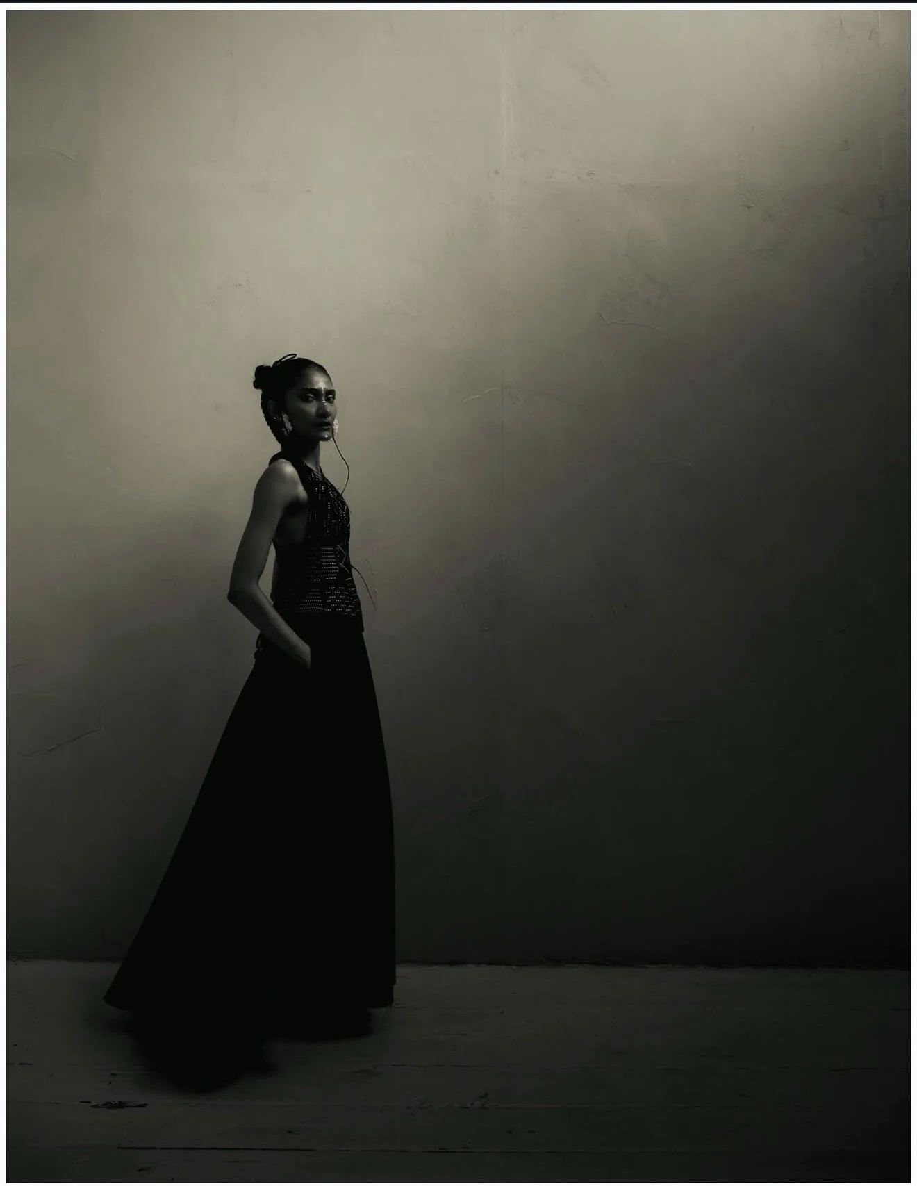 A woman with braided hair and a bun, wearing a sleeveless top and long wide-leg pants, stands against a textured wall with her hands in her pockets, looking to the side in a monochrome setting.