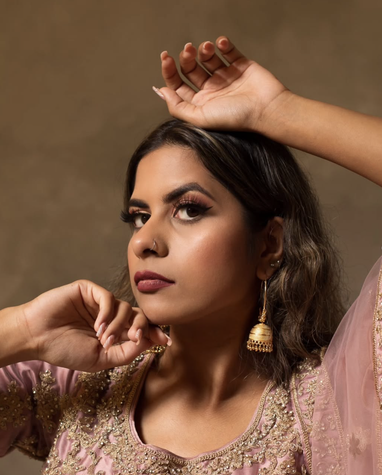 Portrait of a woman with dark hair styled in loose waves, wearing traditional pink and gold embroidered attire, gold earrings, and makeup with bold eyeliner and lipstick. Moveable props and modular set design inside The Space MCR studio in Manchester