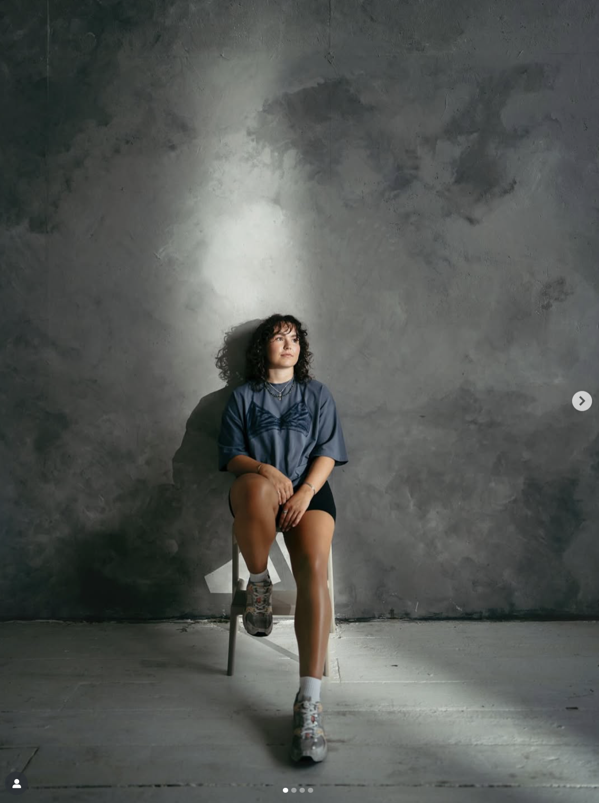 A woman with curly hair sitting on a chair against a textured grey wall, wearing a blue shirt, black shorts, white socks, and sneakers, with a contemplative expression. Creator filming content inside The Space MCR, a content studio for hire in MCR