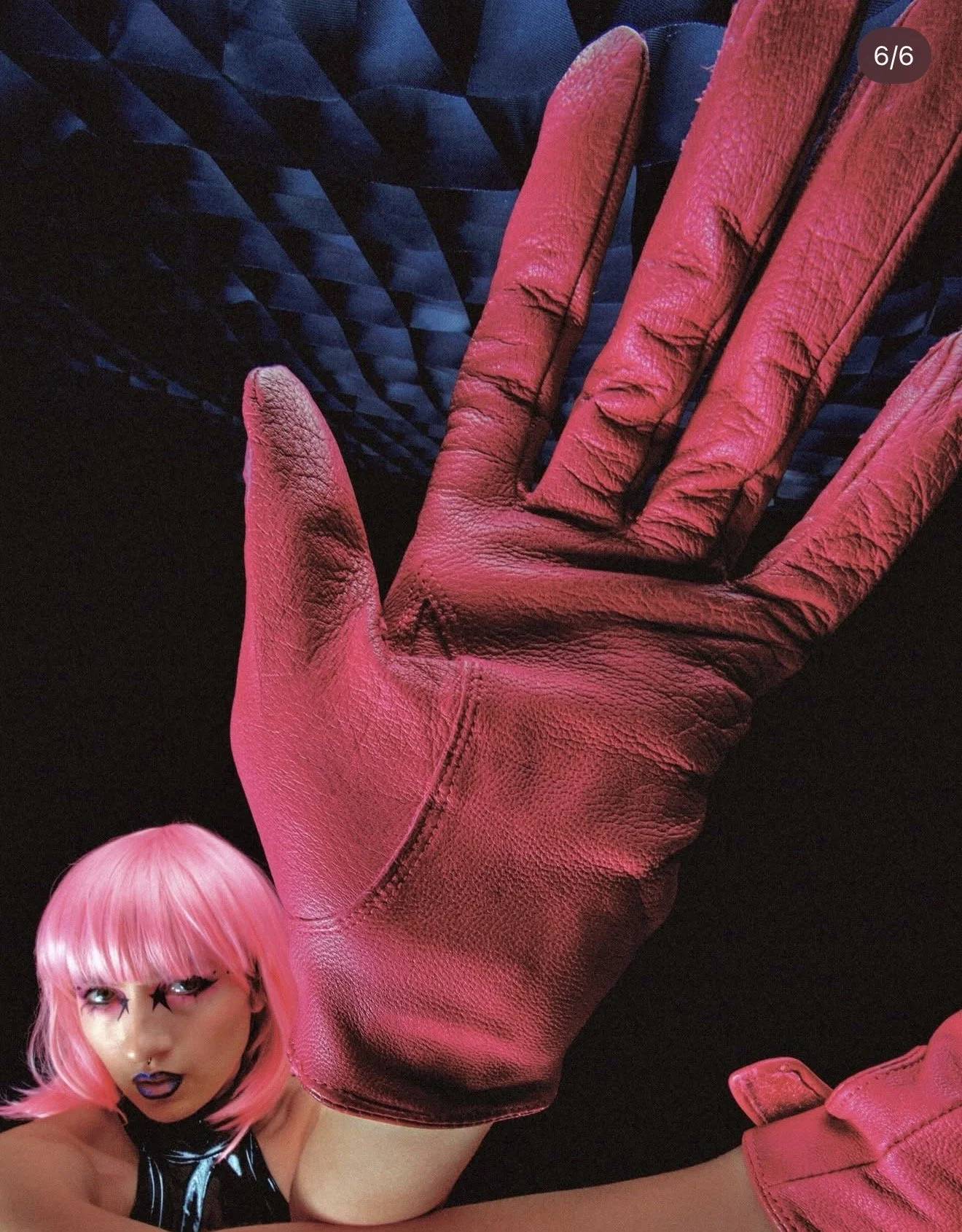 A person with pink hair, makeup, and a shiny black outfit, wearing bright pink leather gloves, with a large pink leather glove in the foreground and a textured blue background.