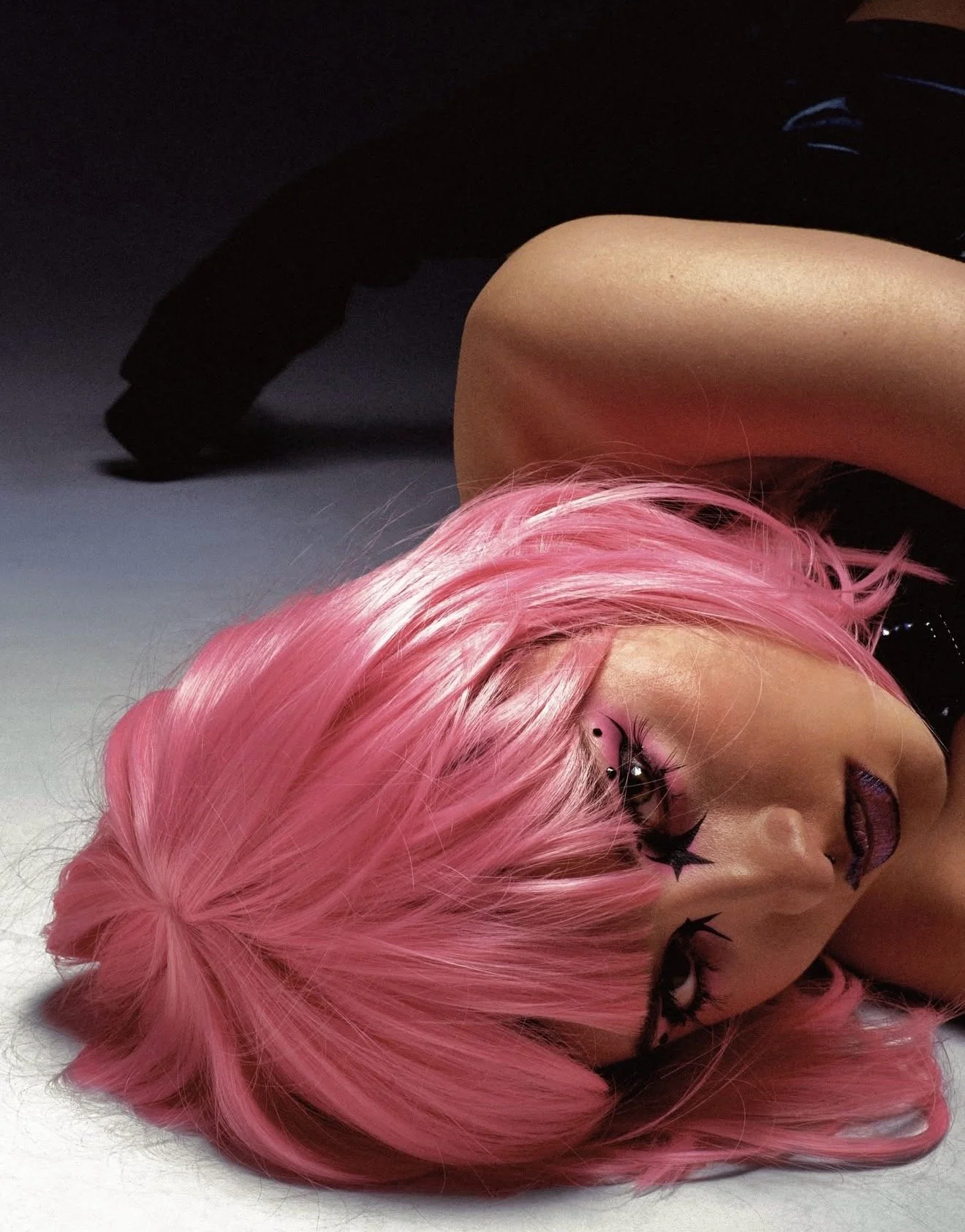 A woman with pink hair, dark makeup, and star-shaped face tattoos lying on the floor, partially visible in the image.