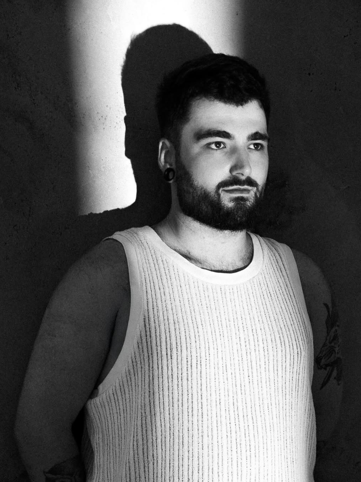 Black and white photo of a young man with a beard and earrings, wearing a sleeveless shirt, standing against a wall with a shadow of his head cast on it. Detail shot of photography set design inside The Space MCR North Manchester studio.