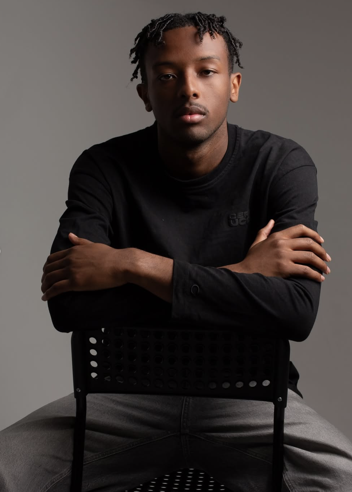 A young man with dreadlocks and medium skin tone sits on a black chair, wearing a black long-sleeve shirt and gray pants, looking directly at the camera against a plain background. Community event inside The Space MCR creative studio – Manchester