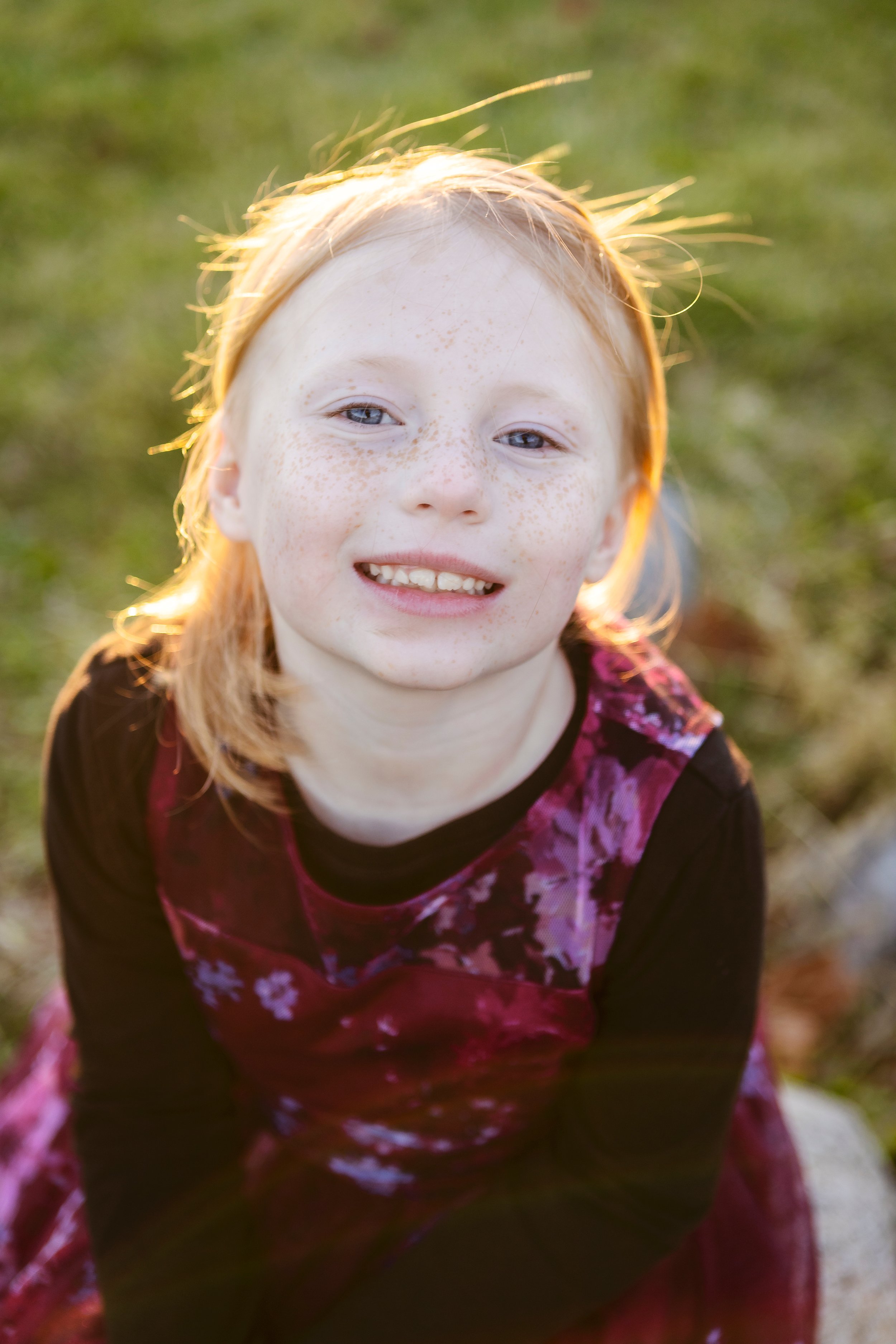 A young girl with red hair and freckles smiling outdoors in sunlight with a blurred grassy background.