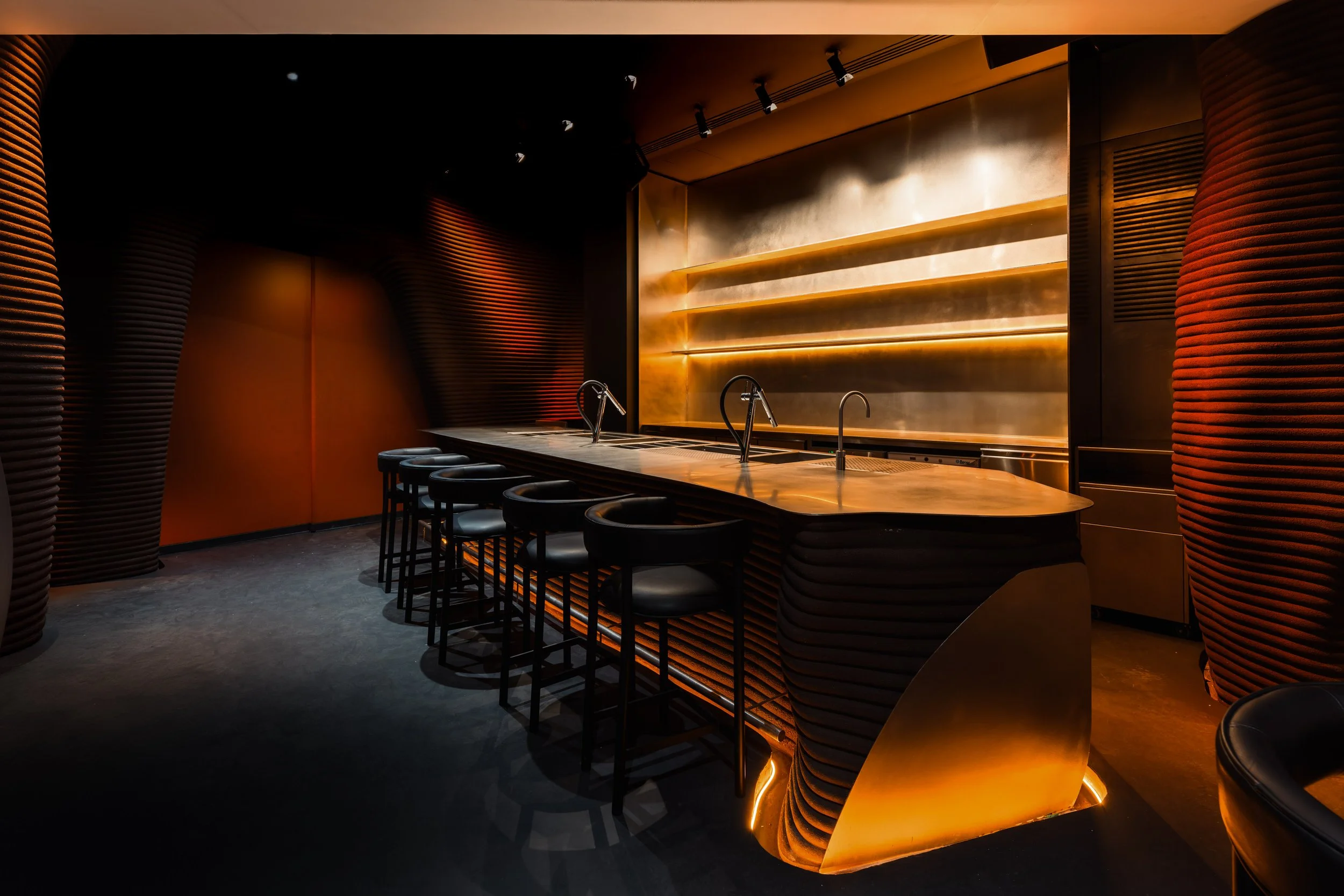 Modern bar with curved walls, illuminated stainless steel bar counter, and four black bar stools.