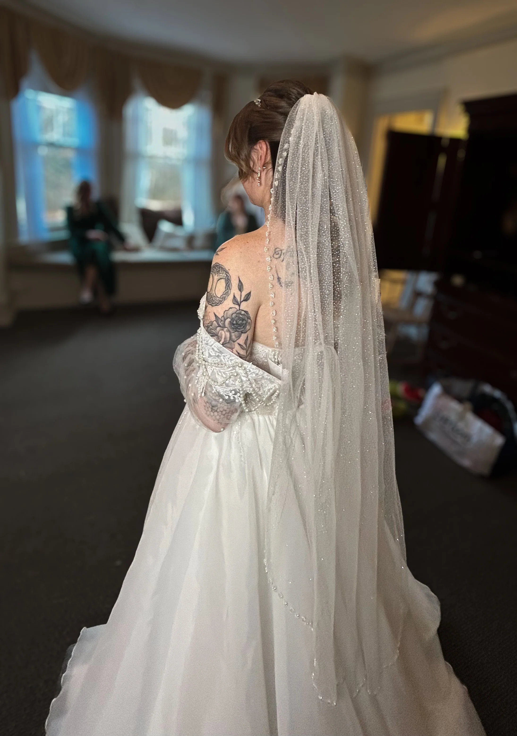 A woman in a wedding dress and veil standing indoors, with tattoos on her arm, while two people are seated on a windowsill in the background.