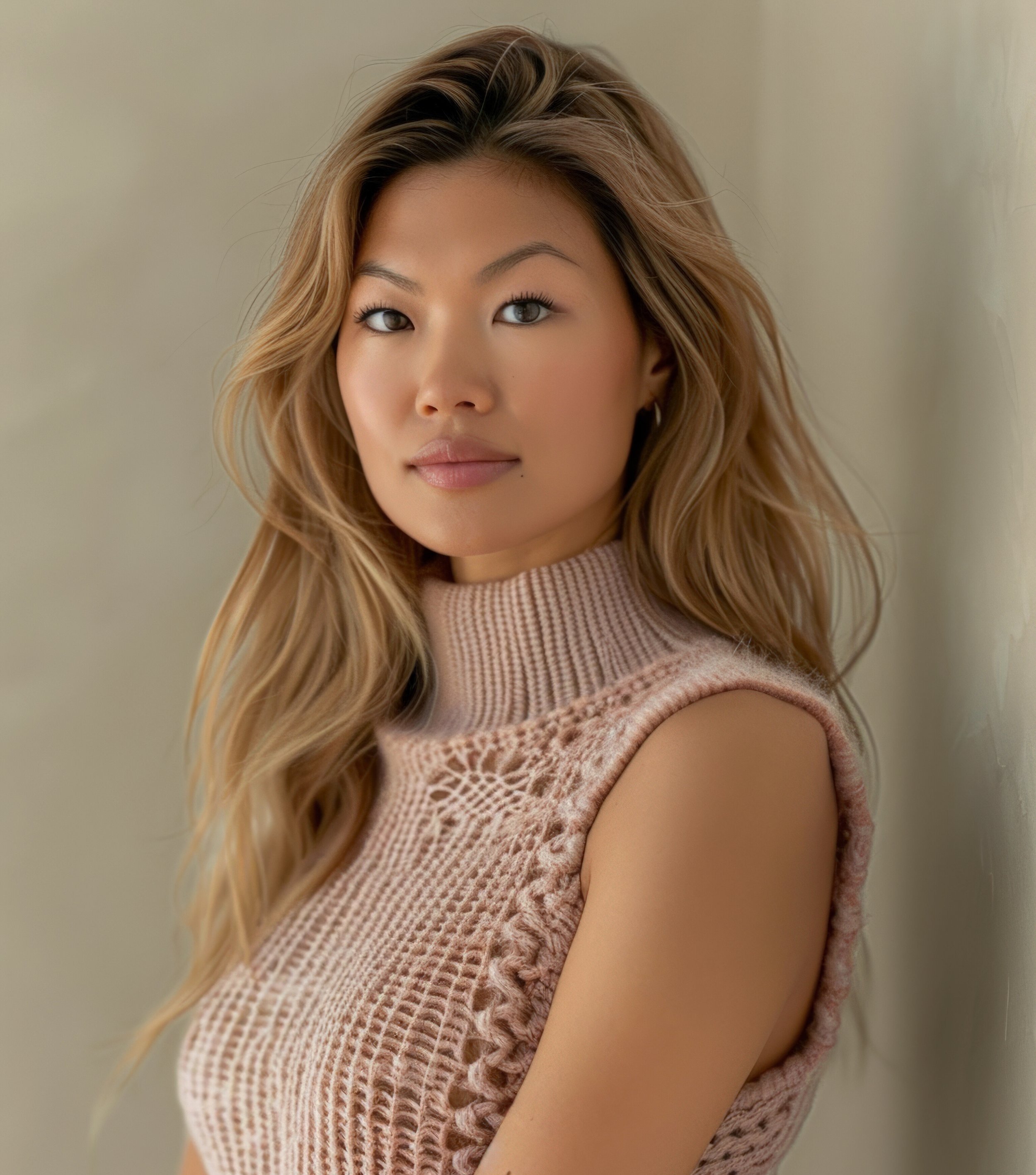 A woman with long, wavy blonde hair and light makeup wearing a sleeveless beige knit top, standing against a neutral background.