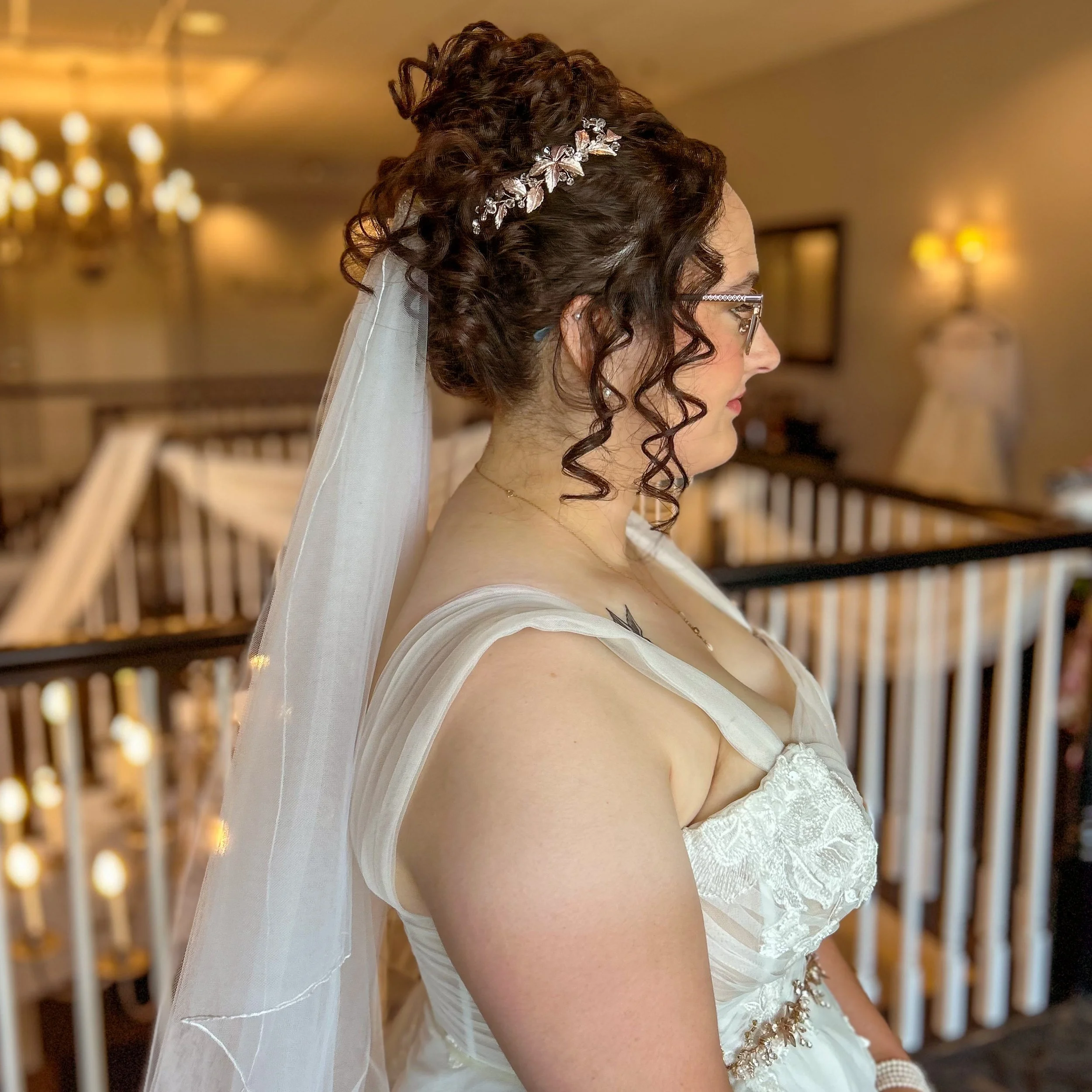 A bride with curly brown hair adorned with a silver hairpiece and veil, wearing glasses and a wedding dress with lace details, standing in a warmly lit venue.