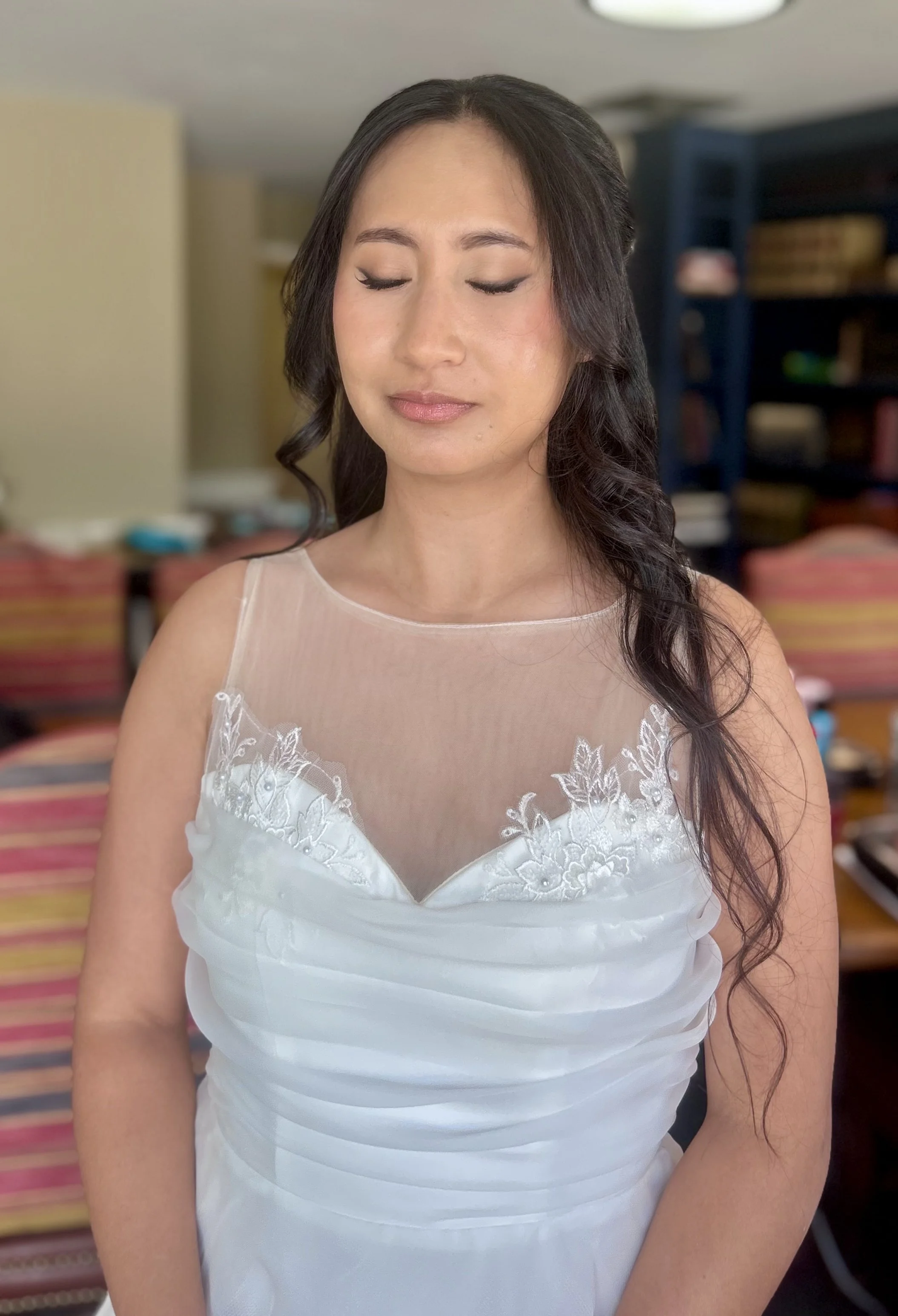A woman in a white wedding dress with lace details, closed eyes, and long wavy dark hair in a room with colorful chairs and a bookshelf.