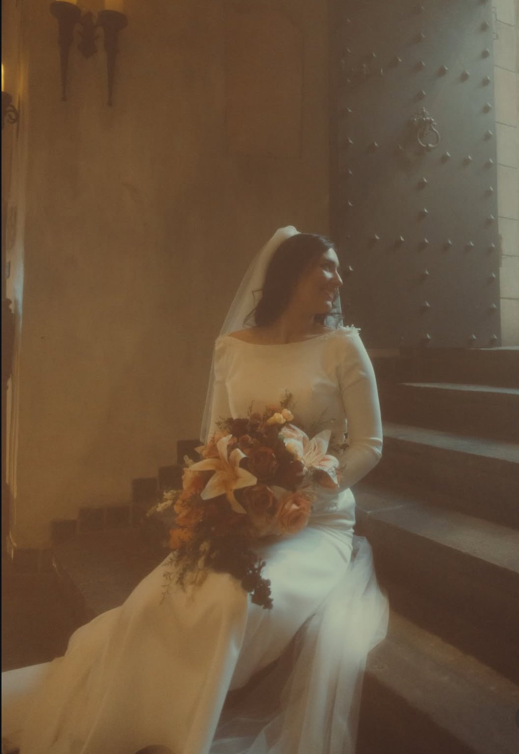 A bride in a white wedding gown sitting on stairs, holding a bouquet of flowers, with a veil on her head, smiling and looking to the side in a warmly lit indoor setting.