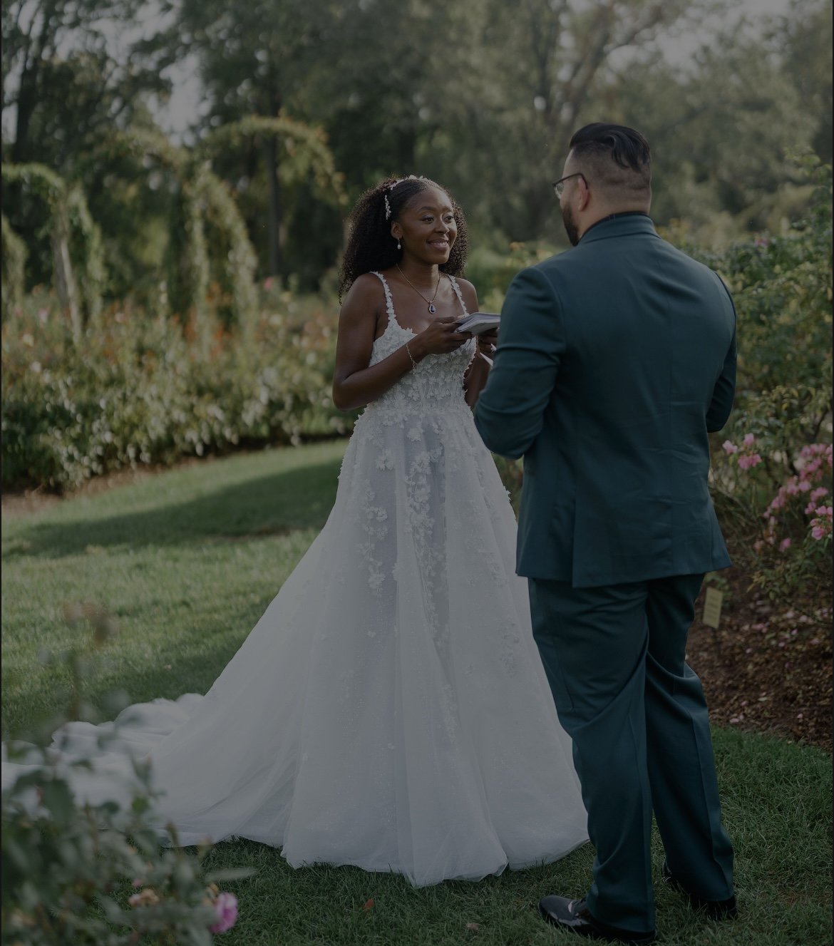 A bride and groom exchanging vows outdoors during daytime, with trees and flowers in the background.