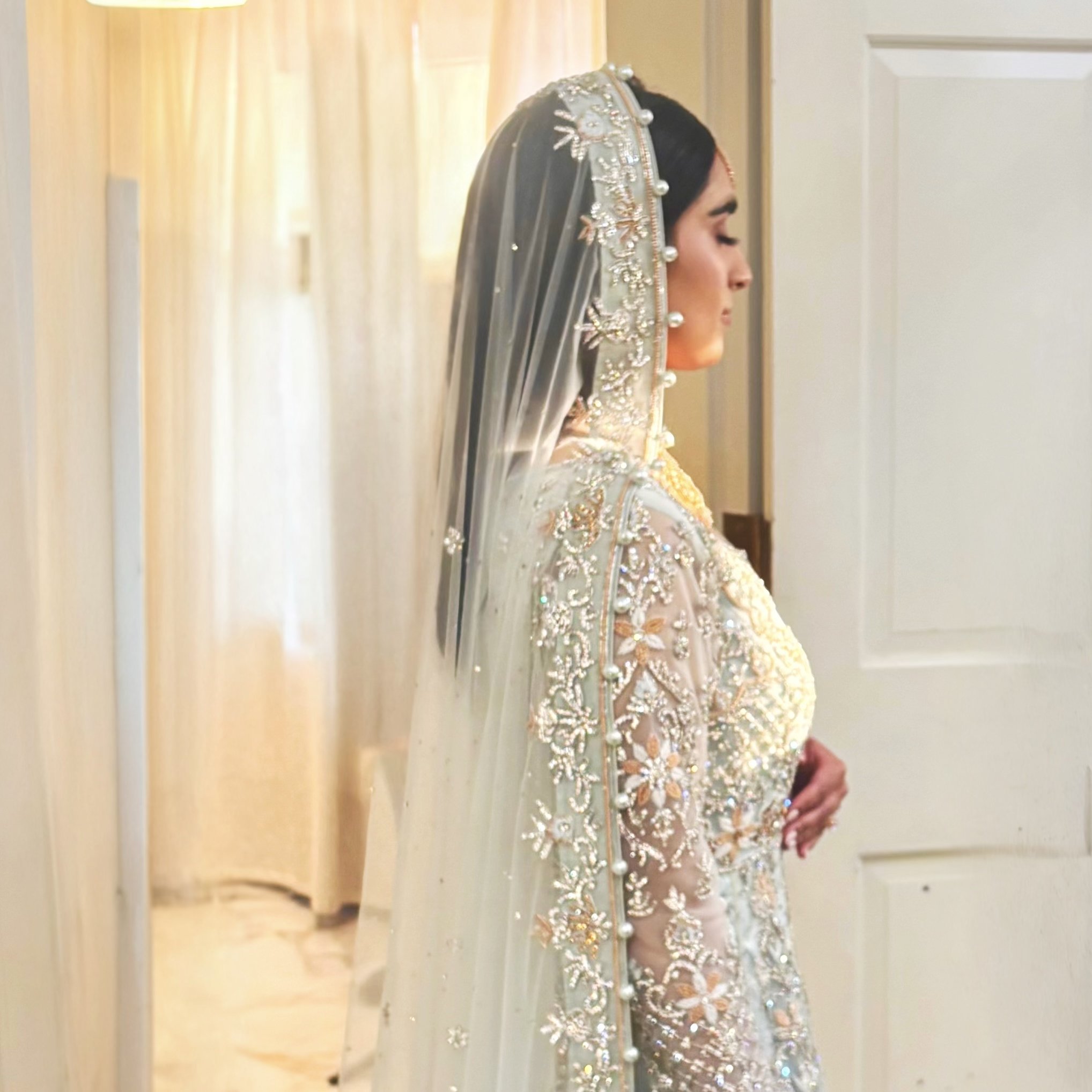 A woman dressed in ornate wedding attire standing indoors with her eyes closed and hands clasped, facing right.