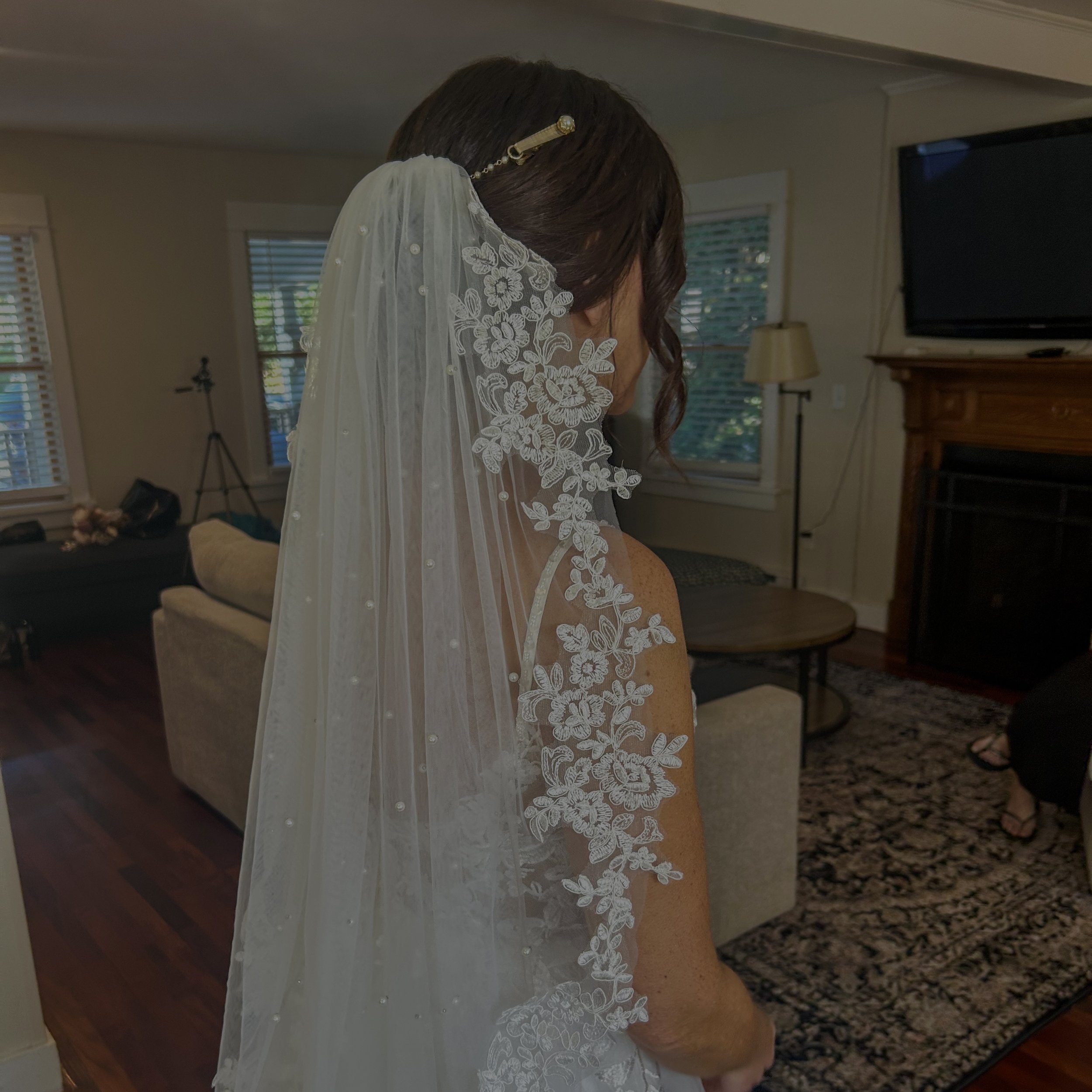 The image shows a woman in a wedding dress with a lace veil. She has dark hair and is standing indoors in a living room with wooden floors, a beige couch, windows with white blinds, a wooden fireplace mantle, and a flat screen TV.