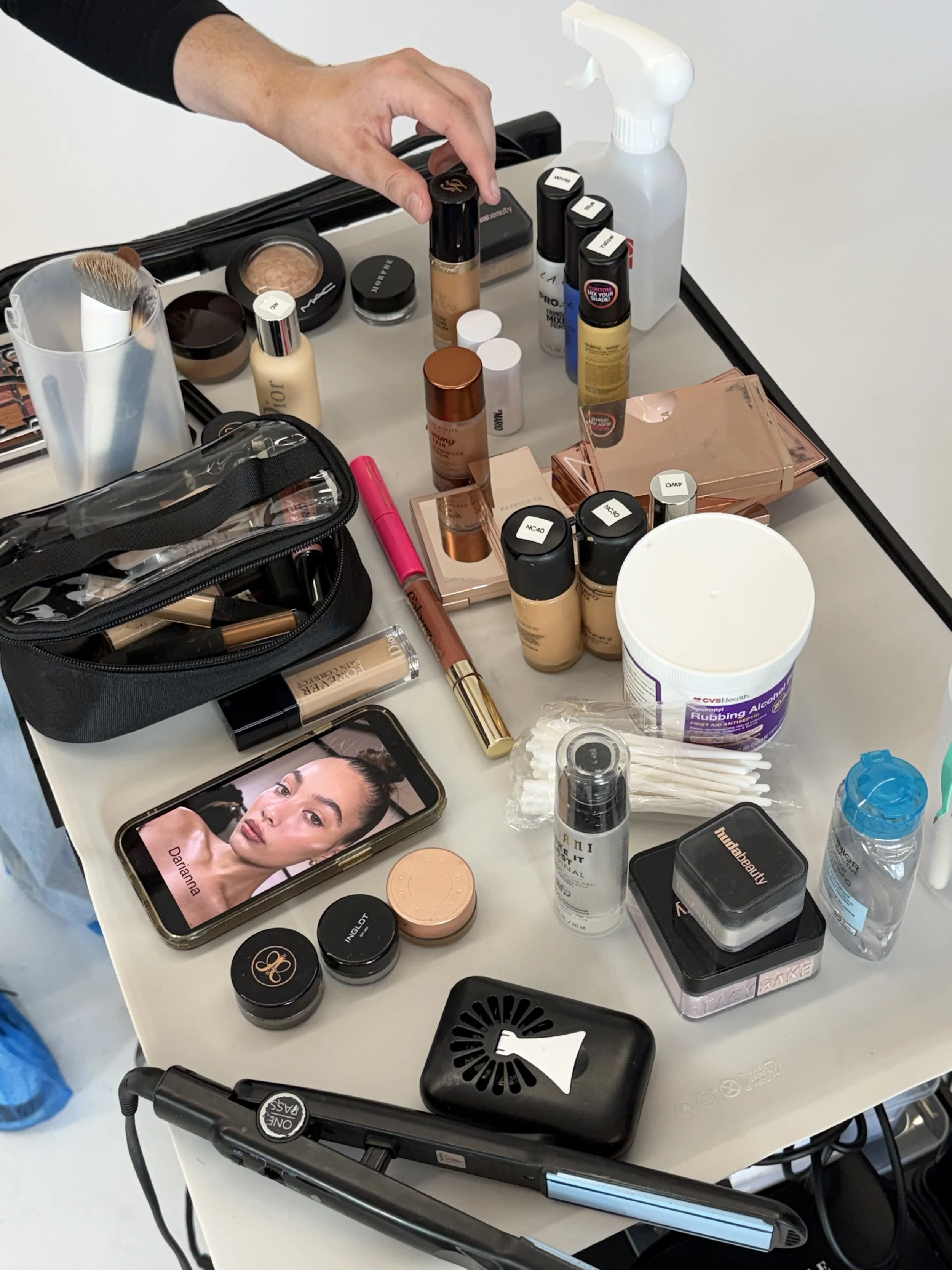 Makeup table with various beauty products, a smartphone showing a woman's face, brushes, foundations, powders, sprays, and other cosmetic items.