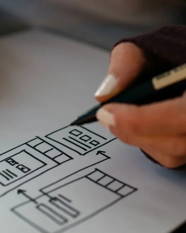 A person's hand with a pencil is drawing a flowchart or diagram on a piece of paper, with arrows and boxes.