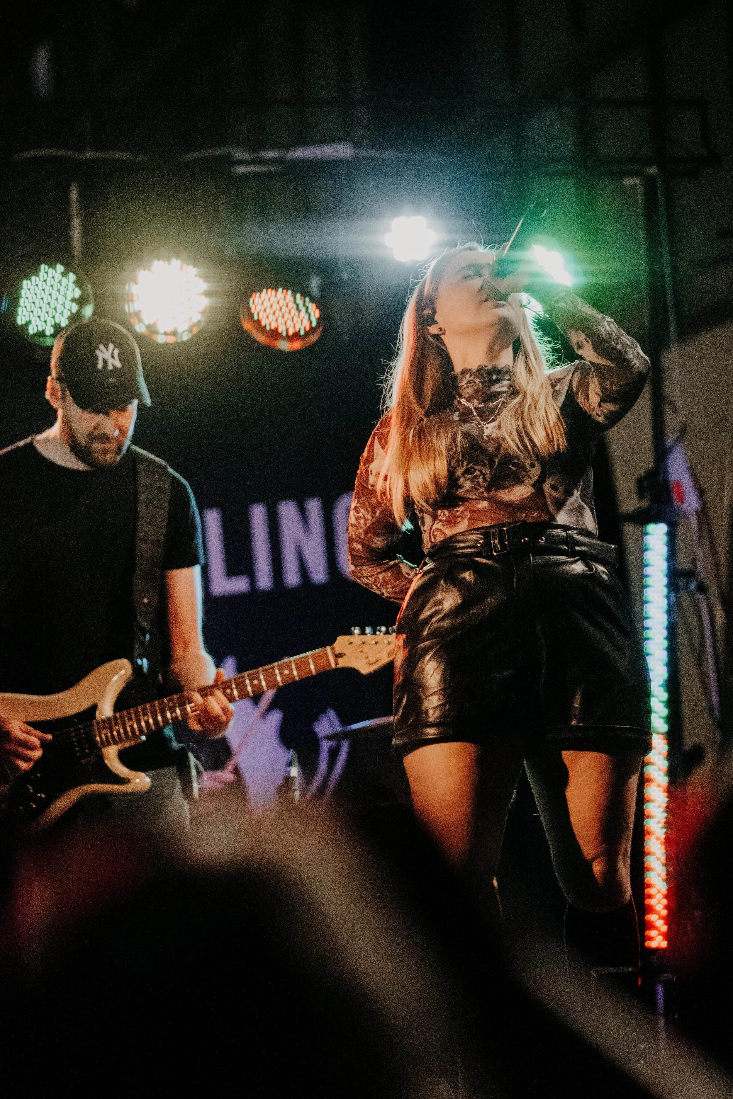Ein Sängerin mit Tattoo-Armen und Lederoutfit singt auf einer Bühne, während ein Gitarrist im schwarzen T-Shirt und Baseballcap im Hintergrund spielt. Bühnenbeleuchtung und ein dunkler Hintergrund sind sichtbar.