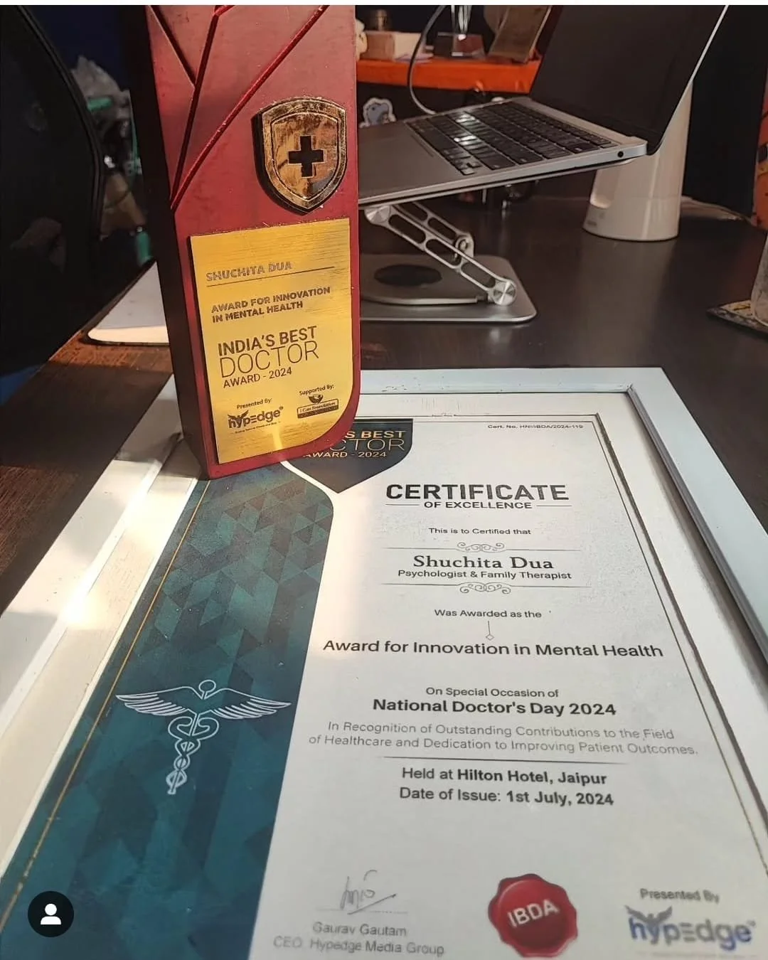 Certificate of excellence awarded to Shuchita Dua, Psychologist & Family Therapist, for innovation in mental health, issued on July 1, 2024, at Hilton Hotel Jaipur. Trophies and office items are also visible.