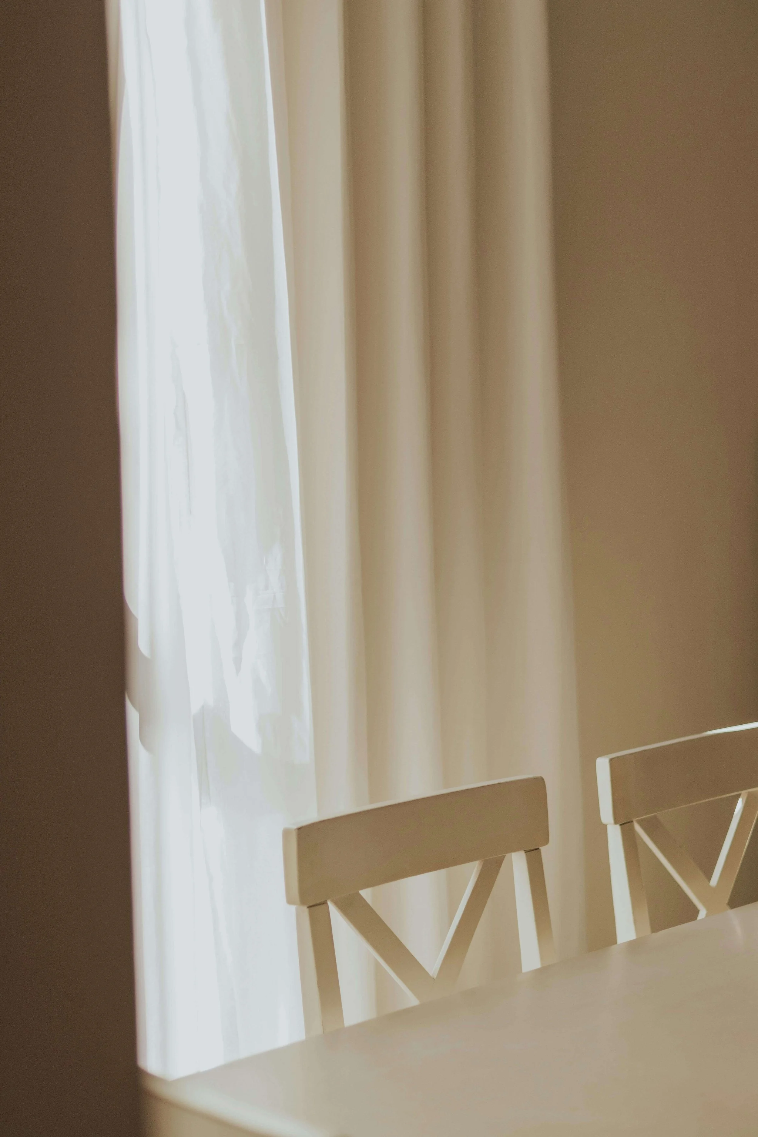 A window with cream-colored curtains allowing natural light into a room. Two white chairs are partially visible in the foreground.