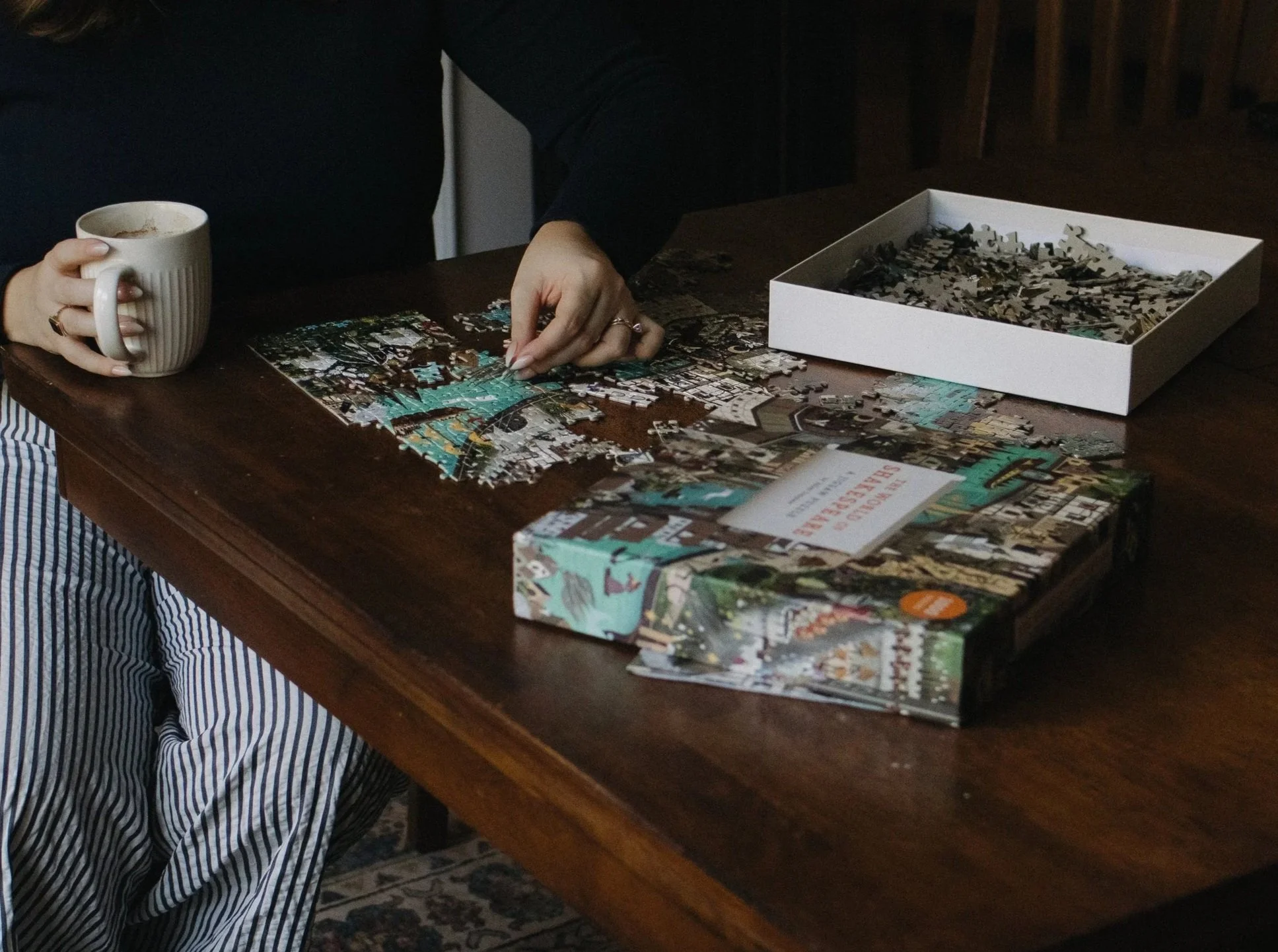 Person assembling a jigsaw puzzle at a wooden table, with a cup in one hand and puzzle pieces on the table and in a box nearby.