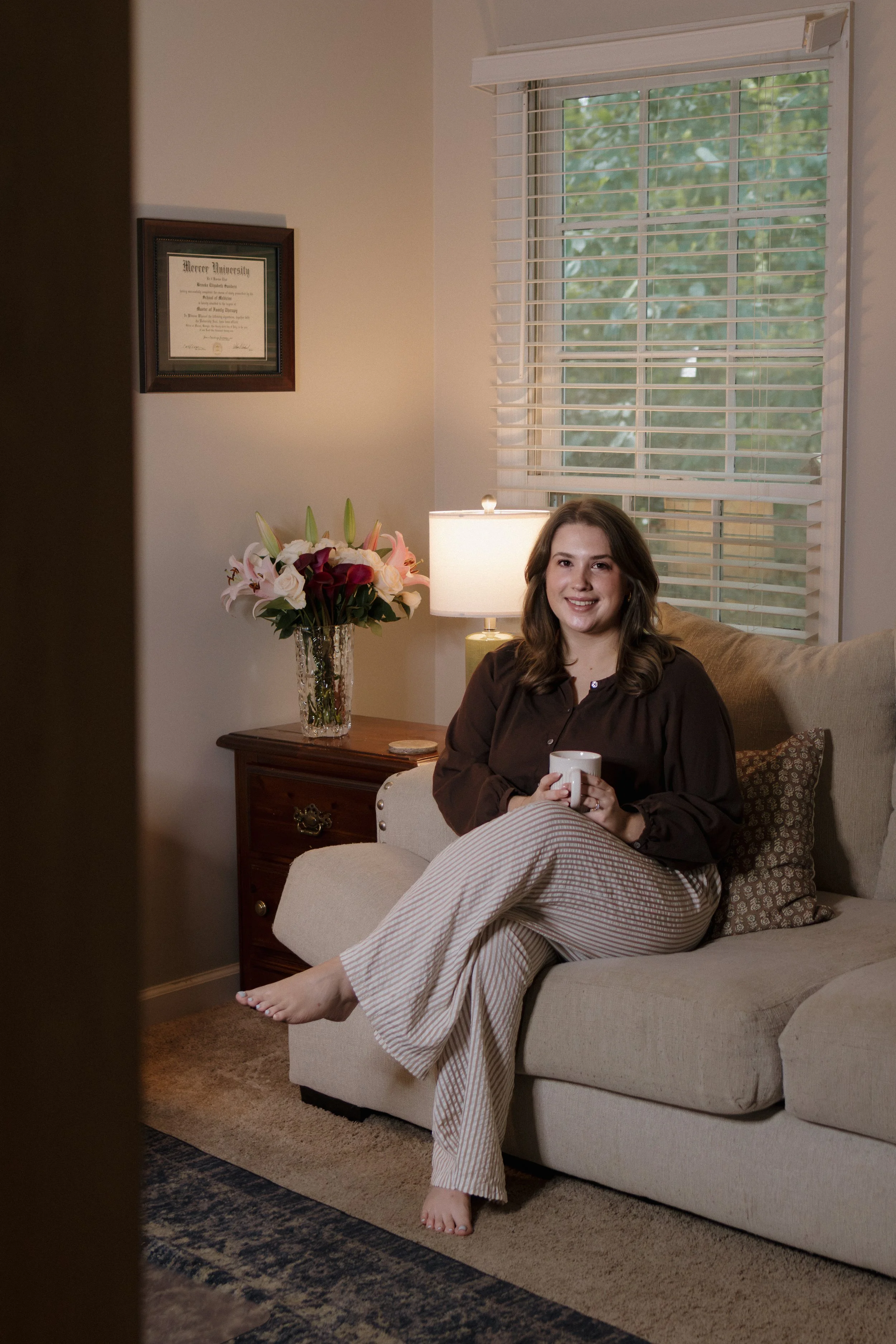 A woman with long brown hair is sitting on a beige couch holding a white mug, smiling at the camera. She is wearing a dark brown top and striped pajama pants. There is a nightstand with a vase of pink and white lilies and a lamp beside her. Behind her, a window with white blinds shows greenery outside. A framed diploma from Mercer University hangs on the wall.