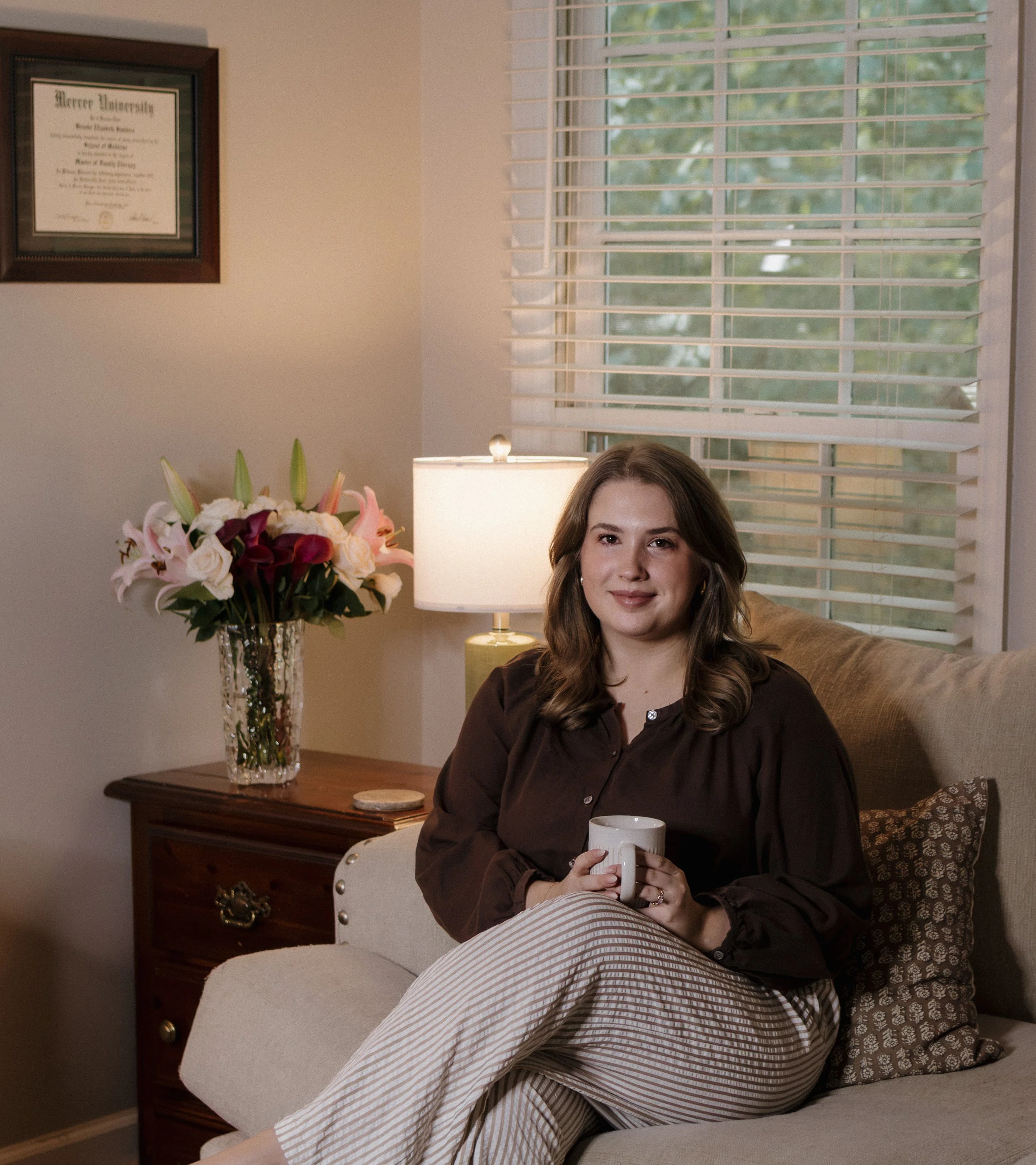 A woman sitting on a beige sofa holding a white mug, with brown hair, wearing a dark brown blouse. Behind her, a window with white blinds showing green trees outside. To her left, a wooden side table with a lamp and a large glass vase of mixed pink, white, and purple flowers. A framed certificate from Mercer University hangs on the wall.