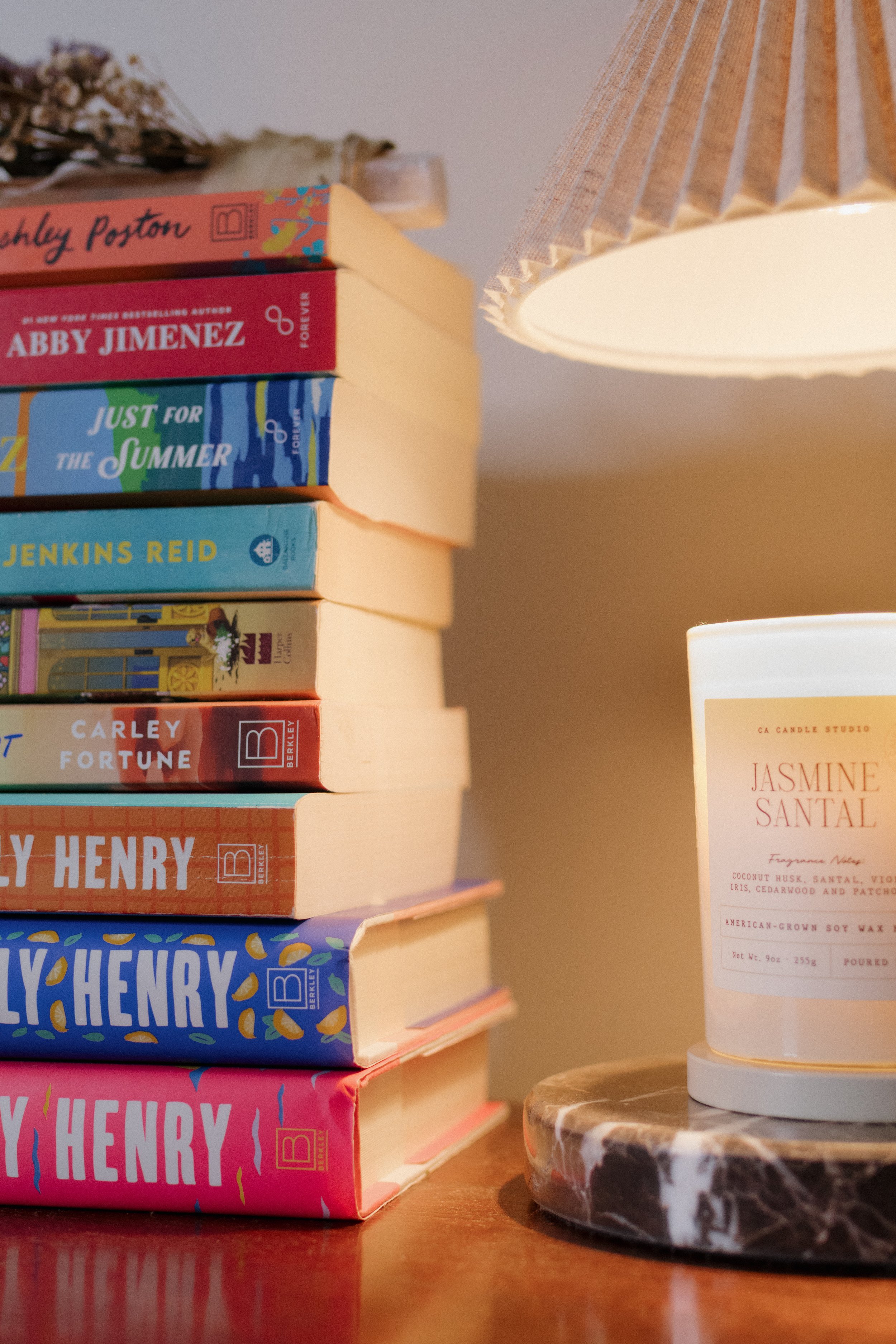 A stack of colorful books with titles by various authors on a wooden surface, beside a lit candle and a marble-patterned coaster. The background includes a table lamp with a pleated shade and some dried flowers.
