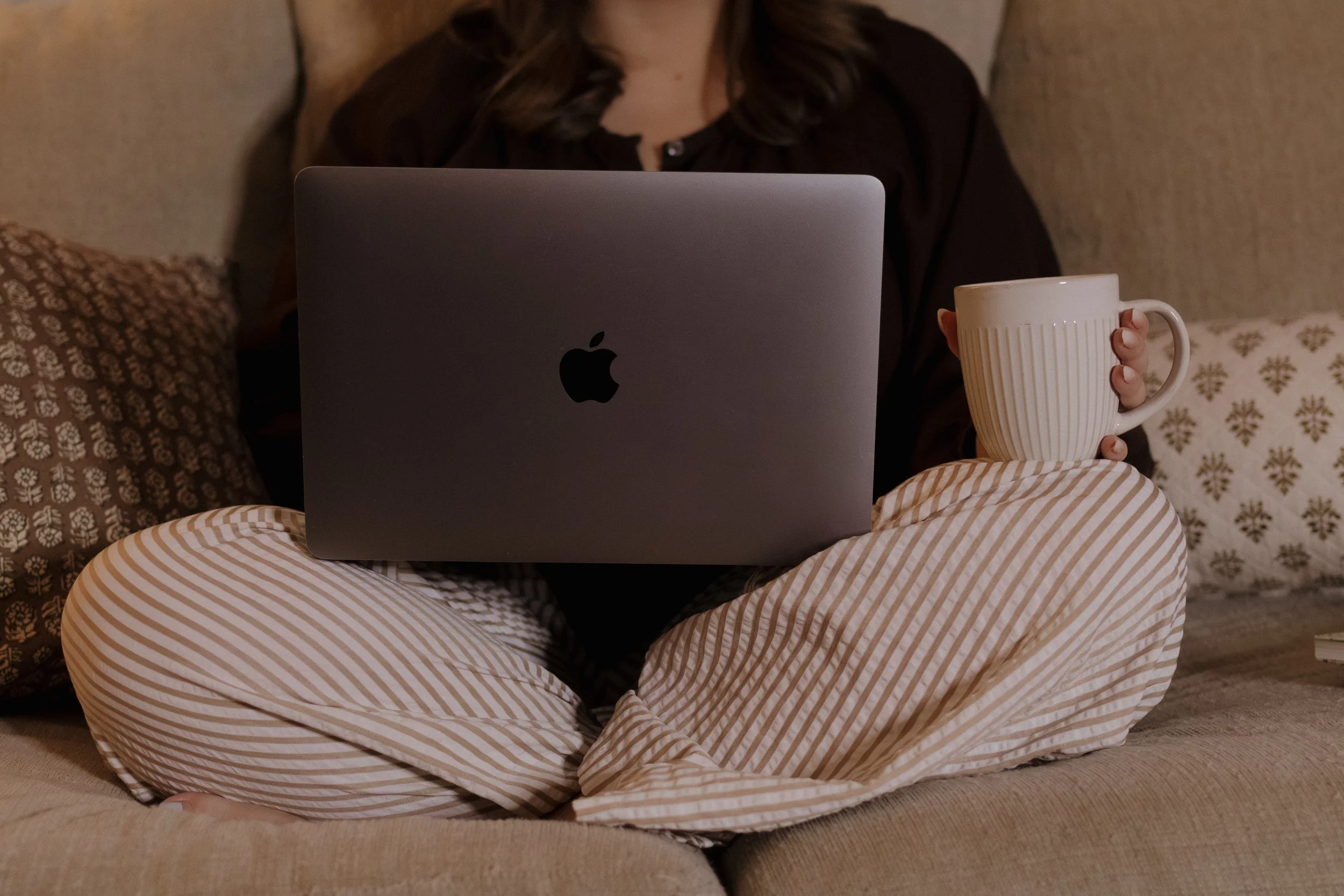 Person sitting on a beige sofa with striped pants, holding a white mug, using a silver MacBook laptop.