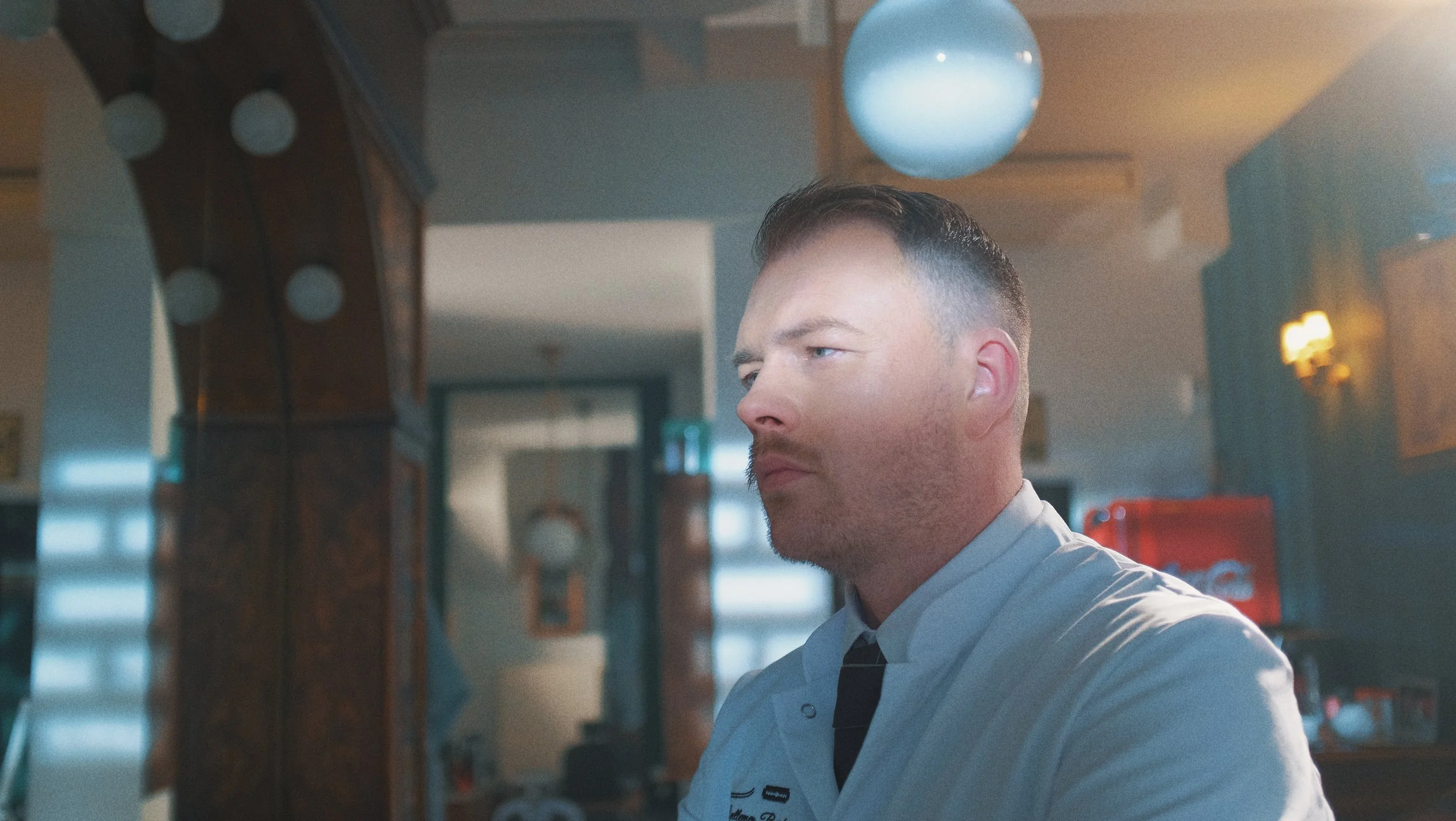 Un homme en uniforme de cuisinier regarde sur le côté dans une cuisine avec des murs en bois, une lumière suspendue et une machine à soda rouge en arrière-plan.