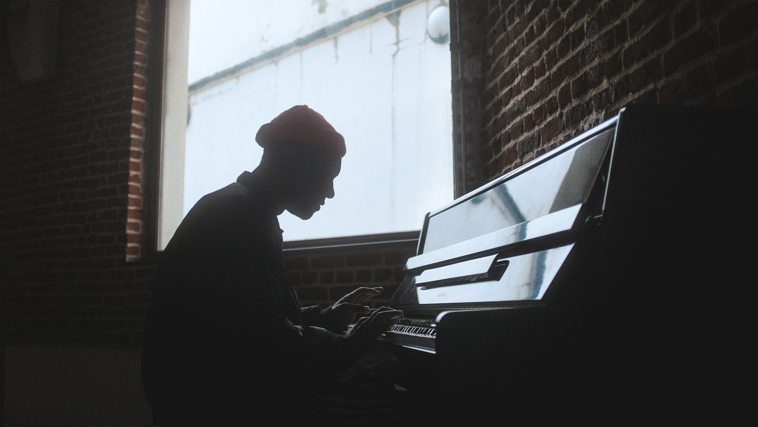 Silhouette d'une personne jouant du piano près d'une fenêtre avec un mur en briques derrière.