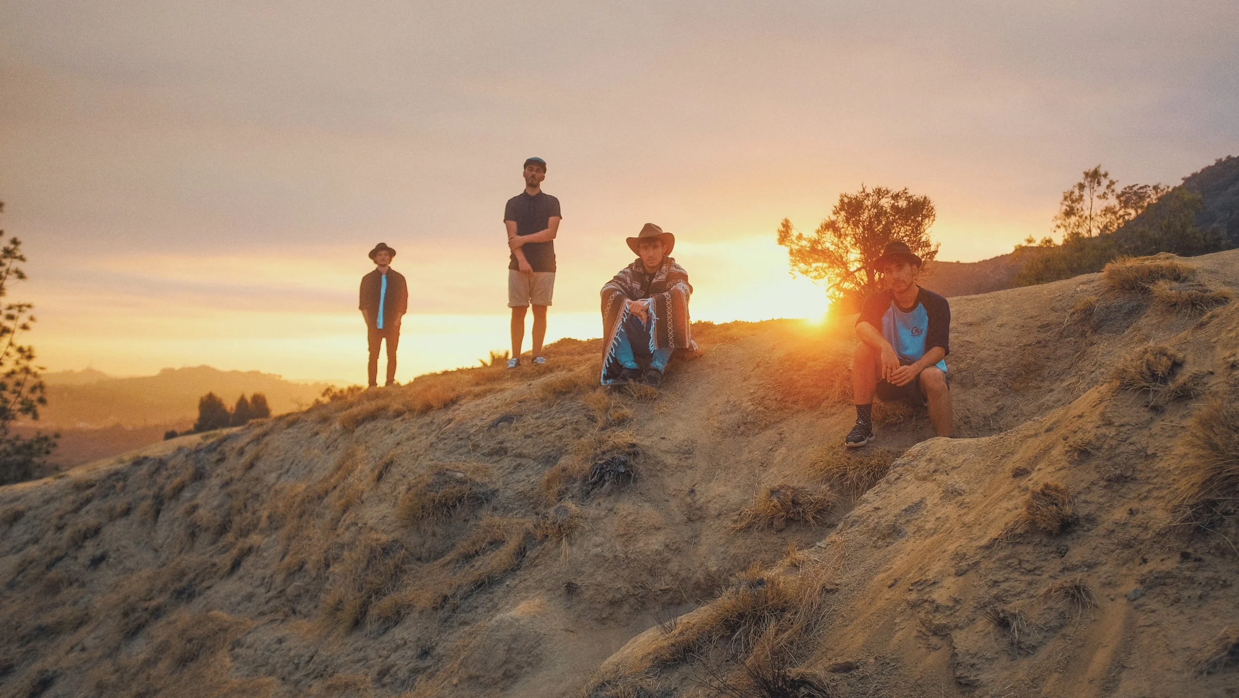 Quatre hommes se tiennent ou s'assoient sur une colline au coucher du soleil, avec un paysage de montagnes en arrière-plan.