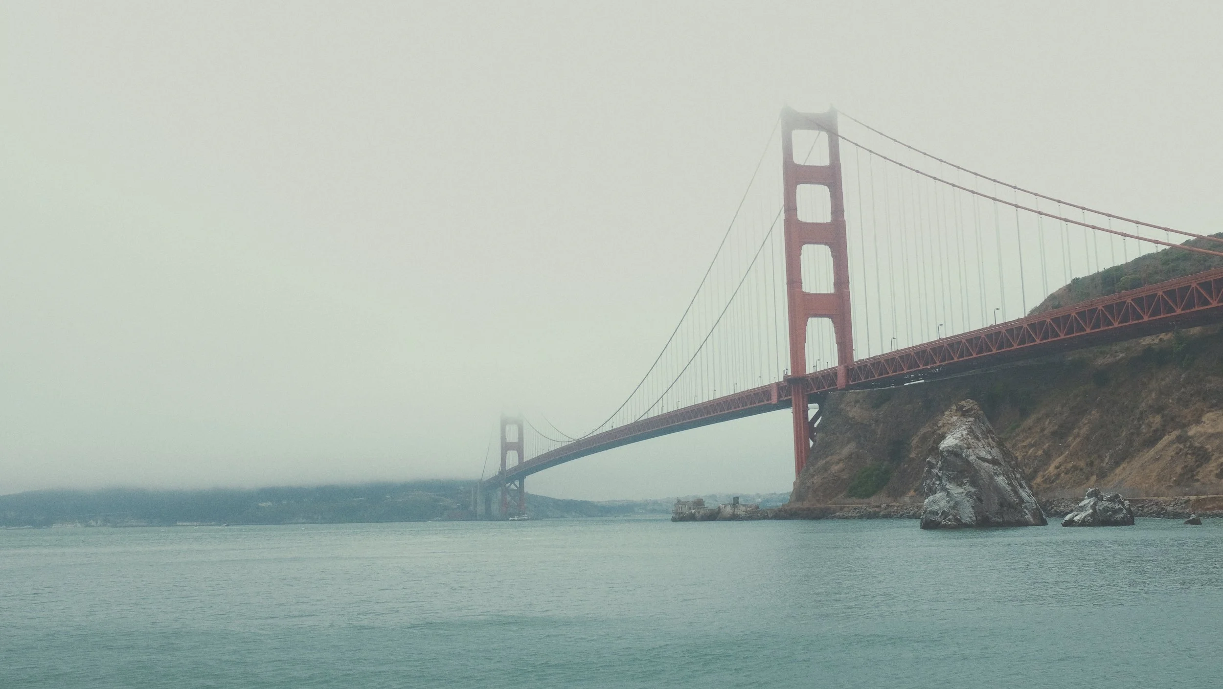 Le pont Golden Gate en Californie sous un ciel brumeux, vu du rivage avec la mer au premier plan et un rocher dans l'eau.