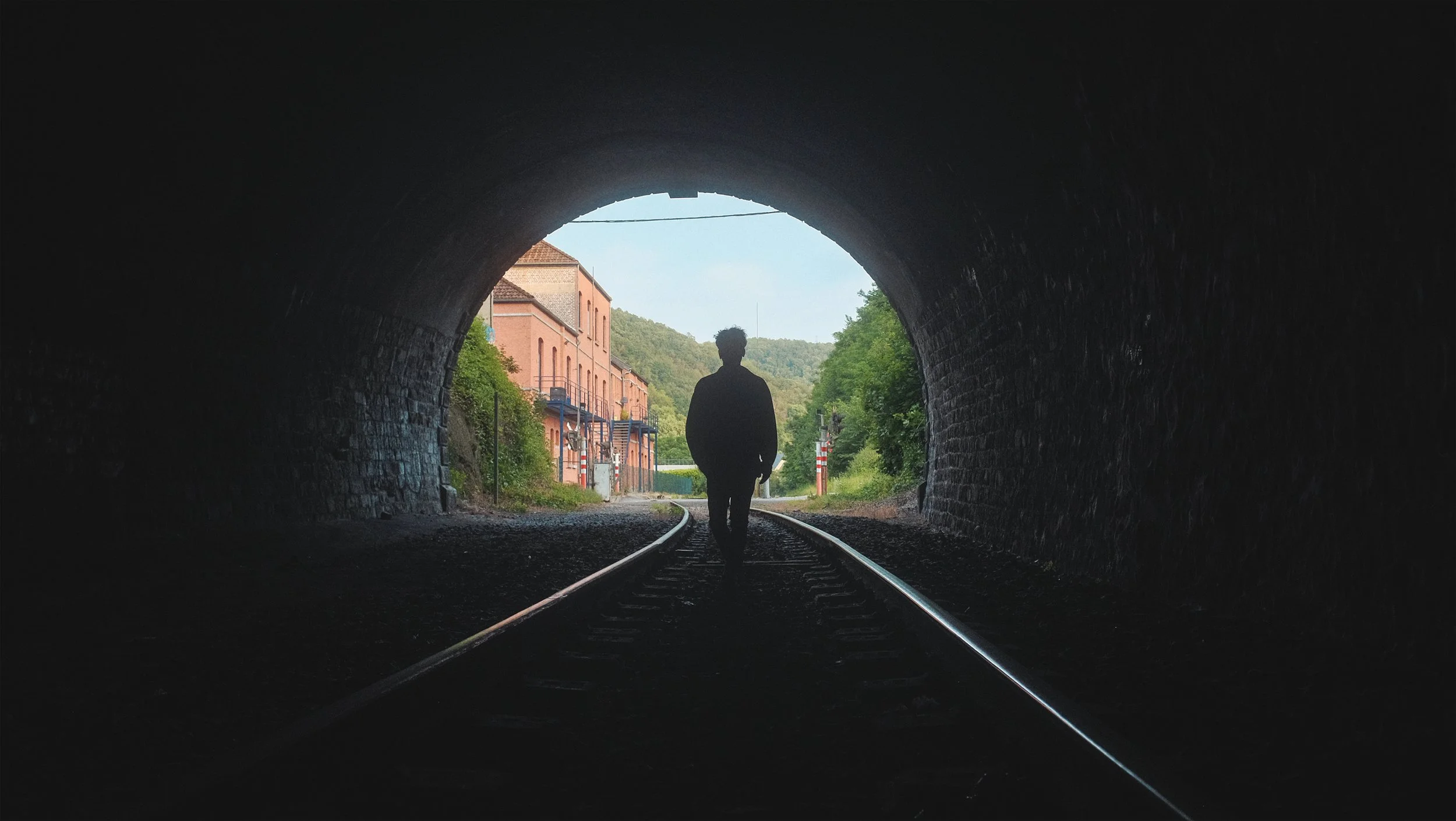 Silhouette d'une personne marchant dans une voie ferrée sous un tunnel, avec un paysage urbain et verdoyant à l'extérieur en arrière-plan.