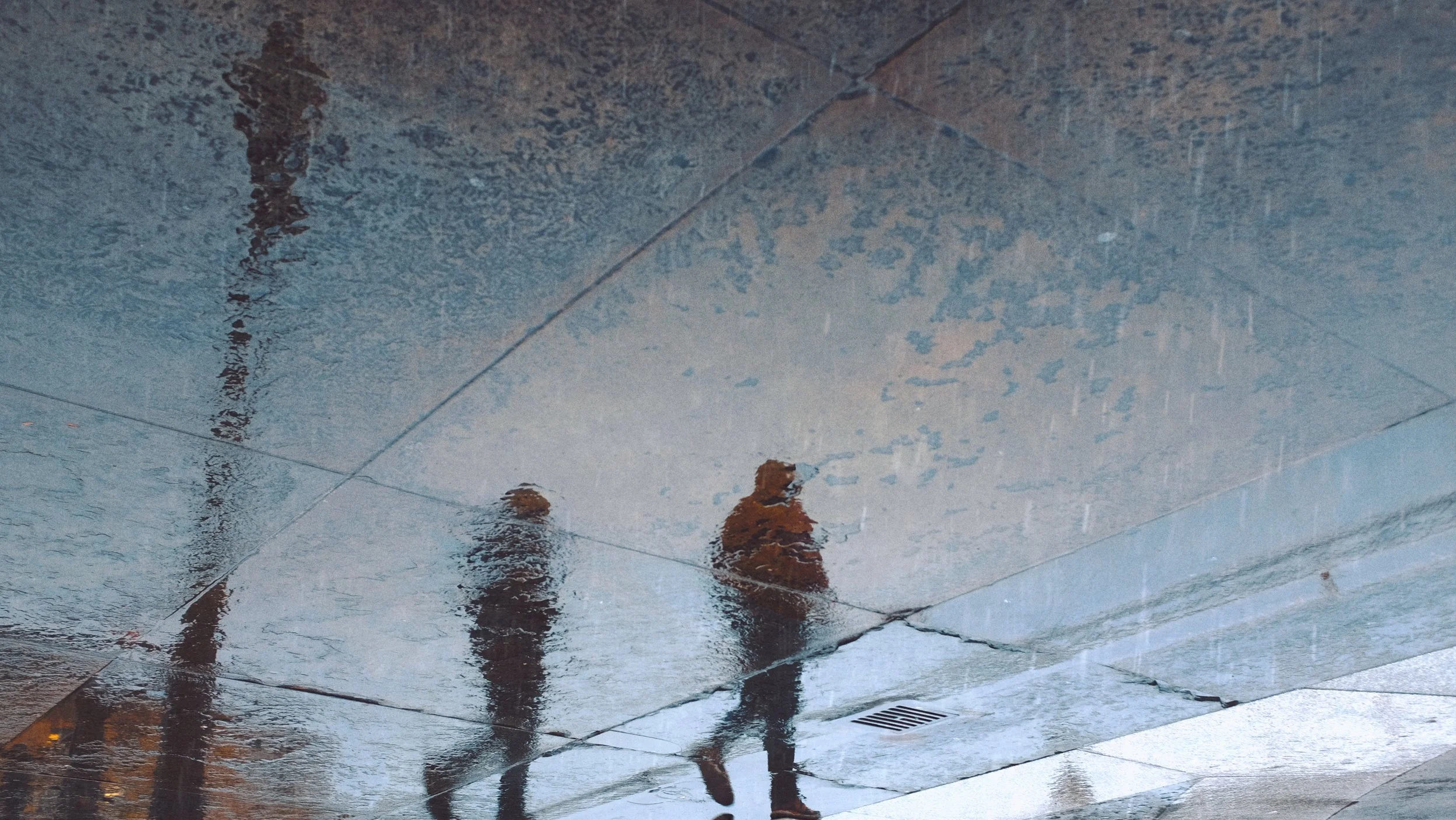 Reflet de deux personnes marchant sous la pluie sur un sol en pierre, avec une partie de la pluie visible dans l'air et des grilles d'évacuation.