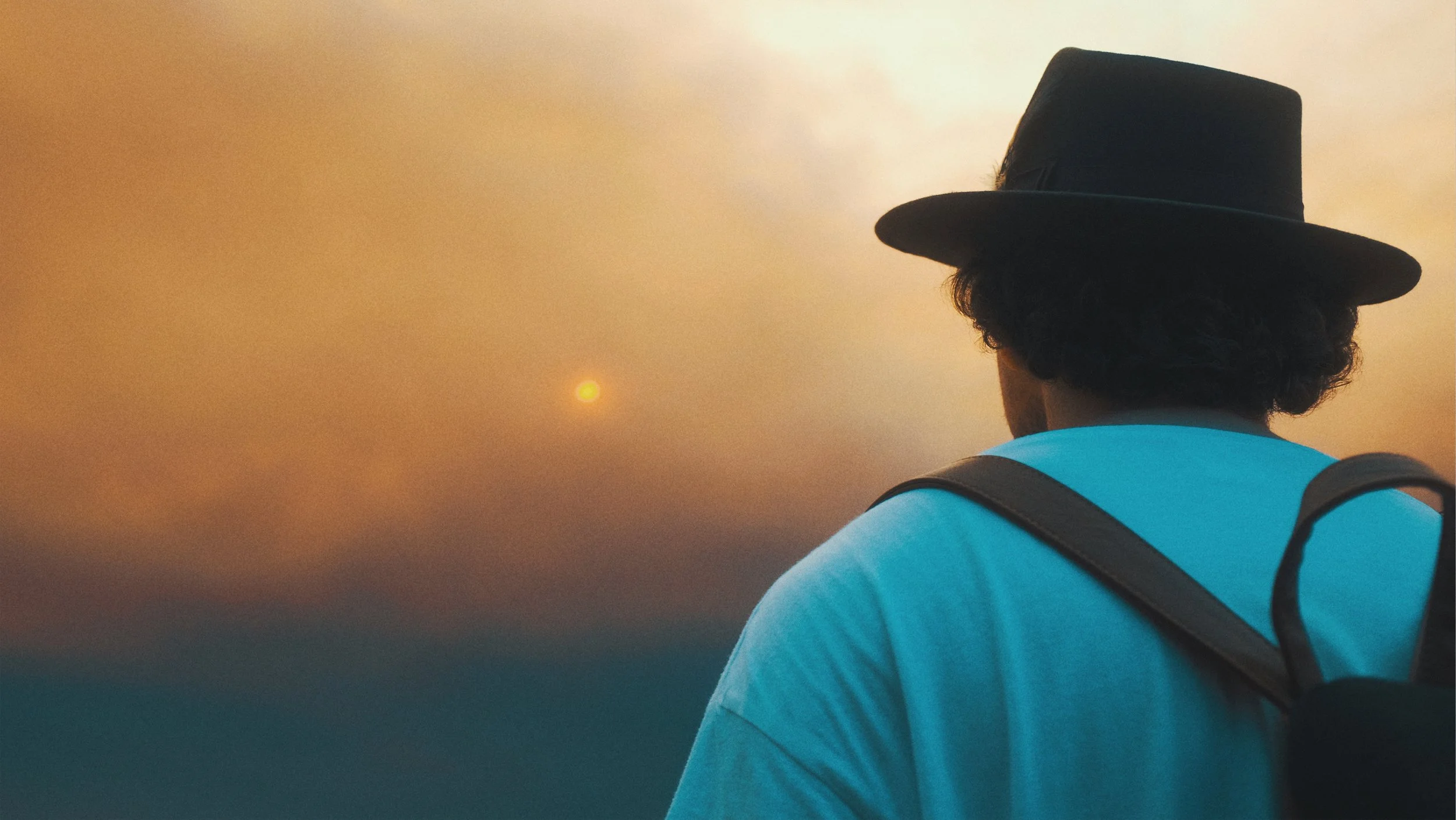Une personne portant un chapeau noir et un t-shirt bleu regarde le soleil couchant, avec une montagne en silhouette en arrière-plan.