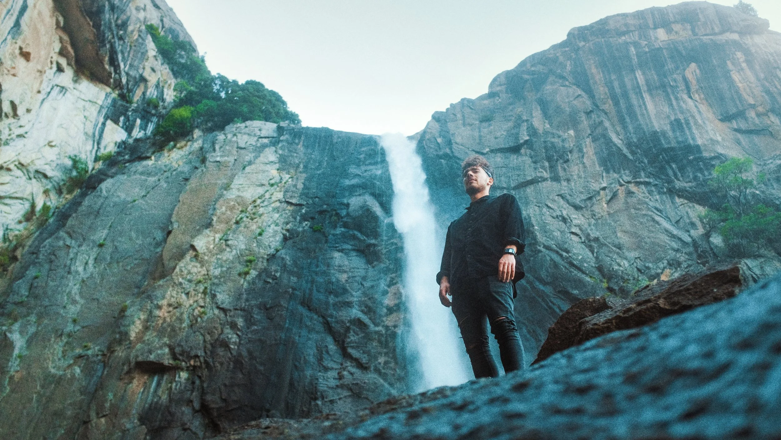 Un homme debout devant une cascade dans un environnement rocheux et verdoyant.