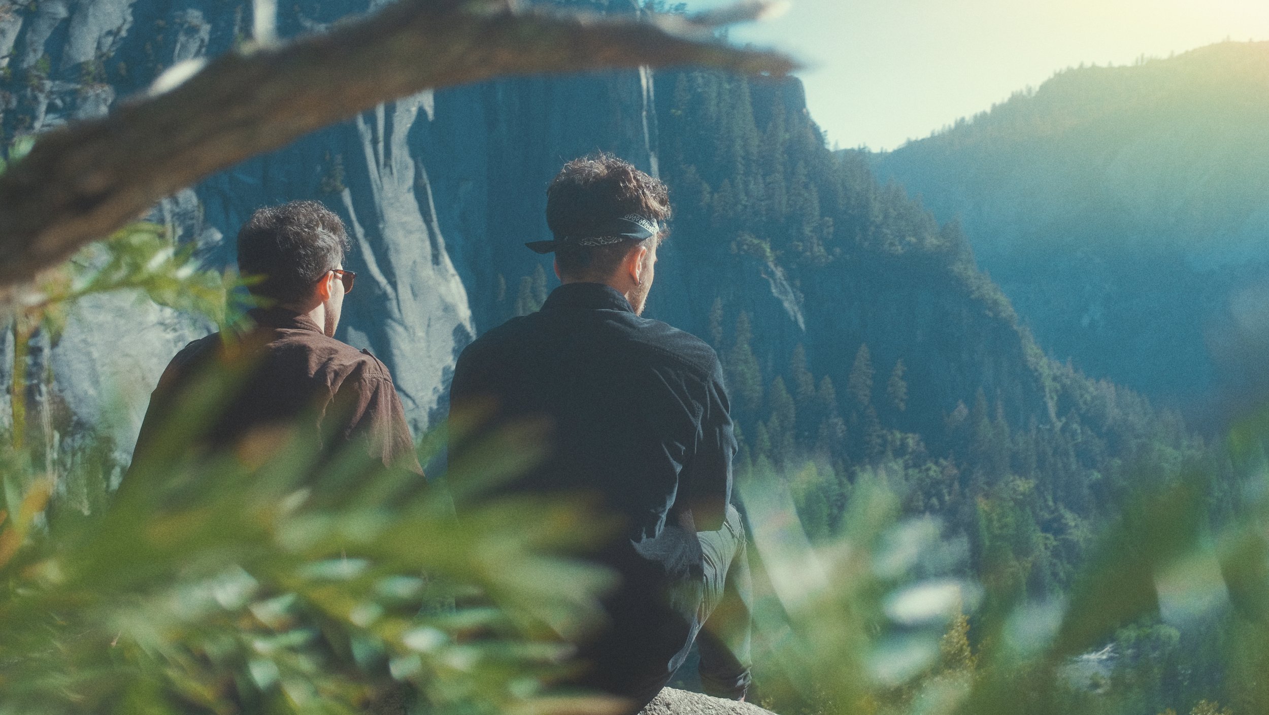 Deux jeunes hommes regardant une vallée verdoyante avec des montagnes en arrière-plan, en position assise, dans un cadre naturel ensoleillé.