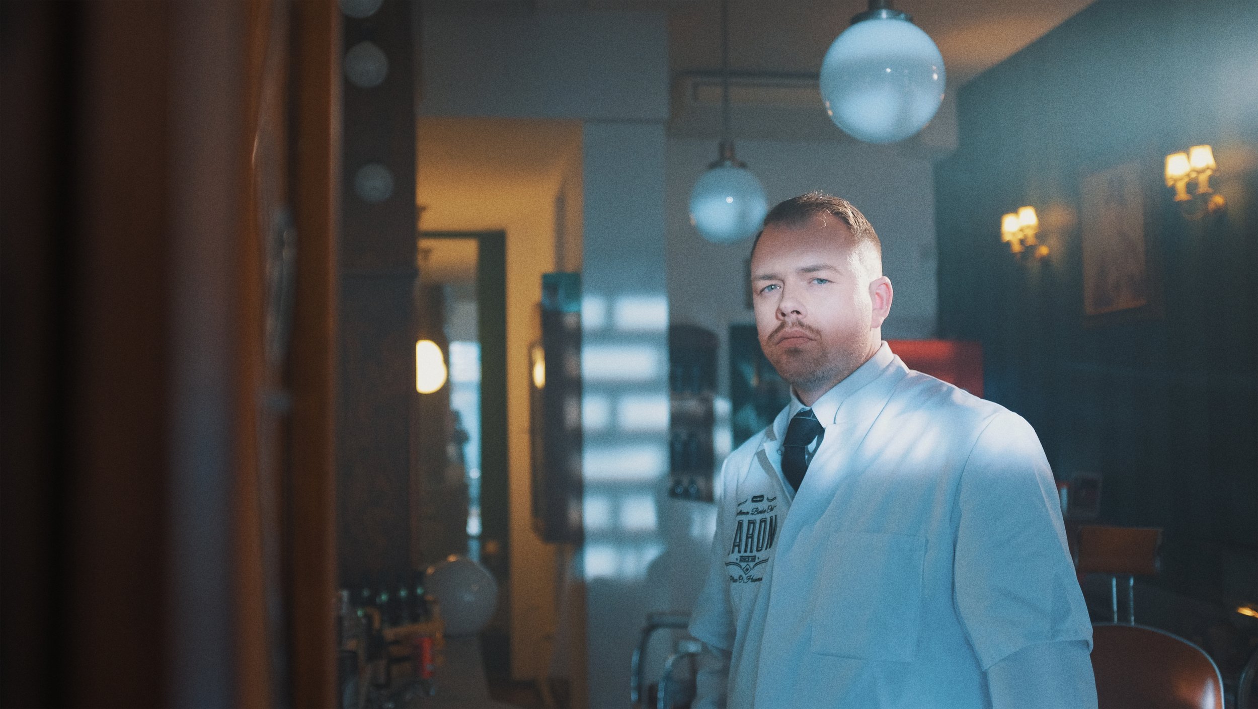 Un homme portant une chemise blanche et une cravate noire dans un salon ou un restaurant, avec un éclairage tamisé et des lampes murales.