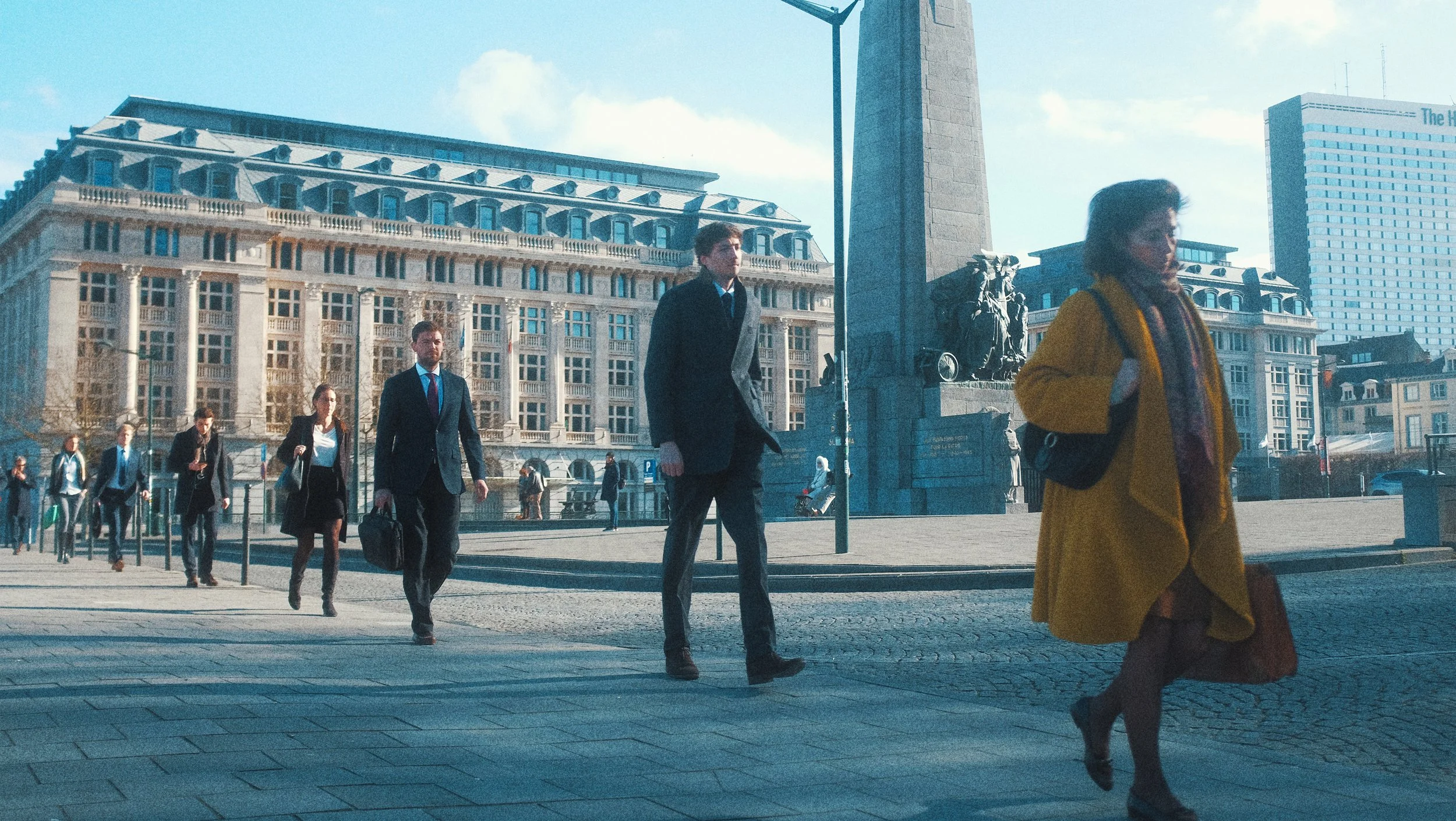Des personnes habillées en vêtements d'affaires traversent une place urbaine avec des bâtiments anciens en arrière-plan. Il y a également une femme en manteau jaune qui marche rapidement en premier plan.